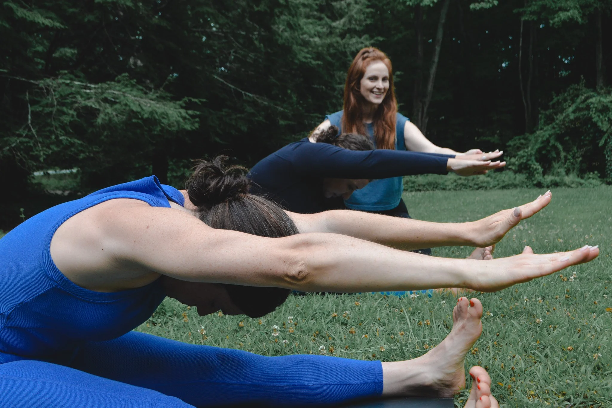 A group of people doing pilates outdoors in a grassy area surrounded by trees, with women doing the Spine Stretch Forward exercise. One woman in blue is reaching forward with her arms extended.