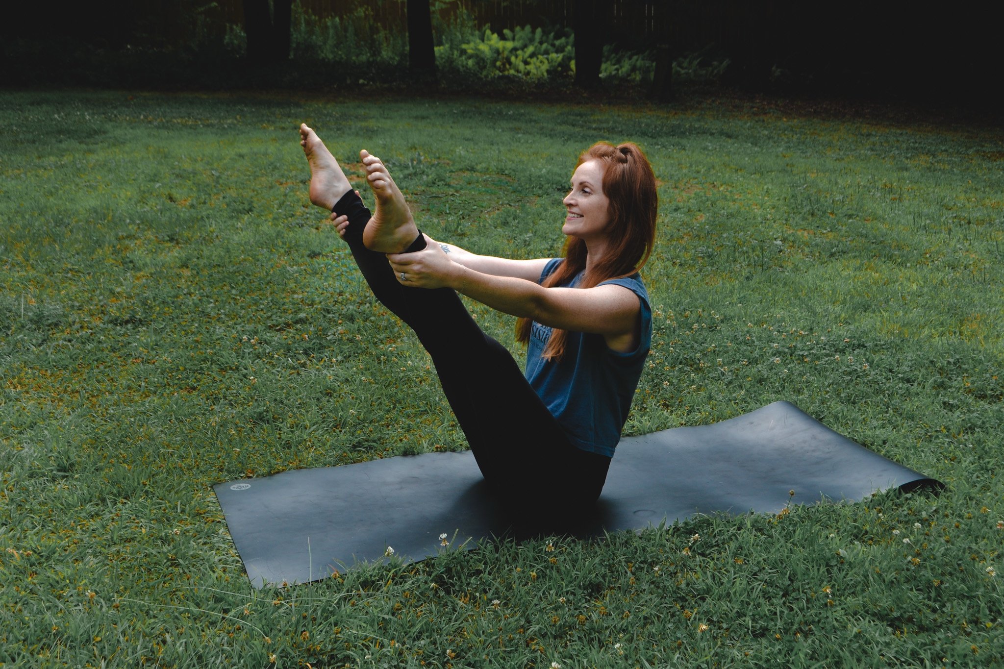 A woman ready for the open leg rocker pilates exercise on a black mat in a green grassy field, holding her legs, smiling, with trees in the background.