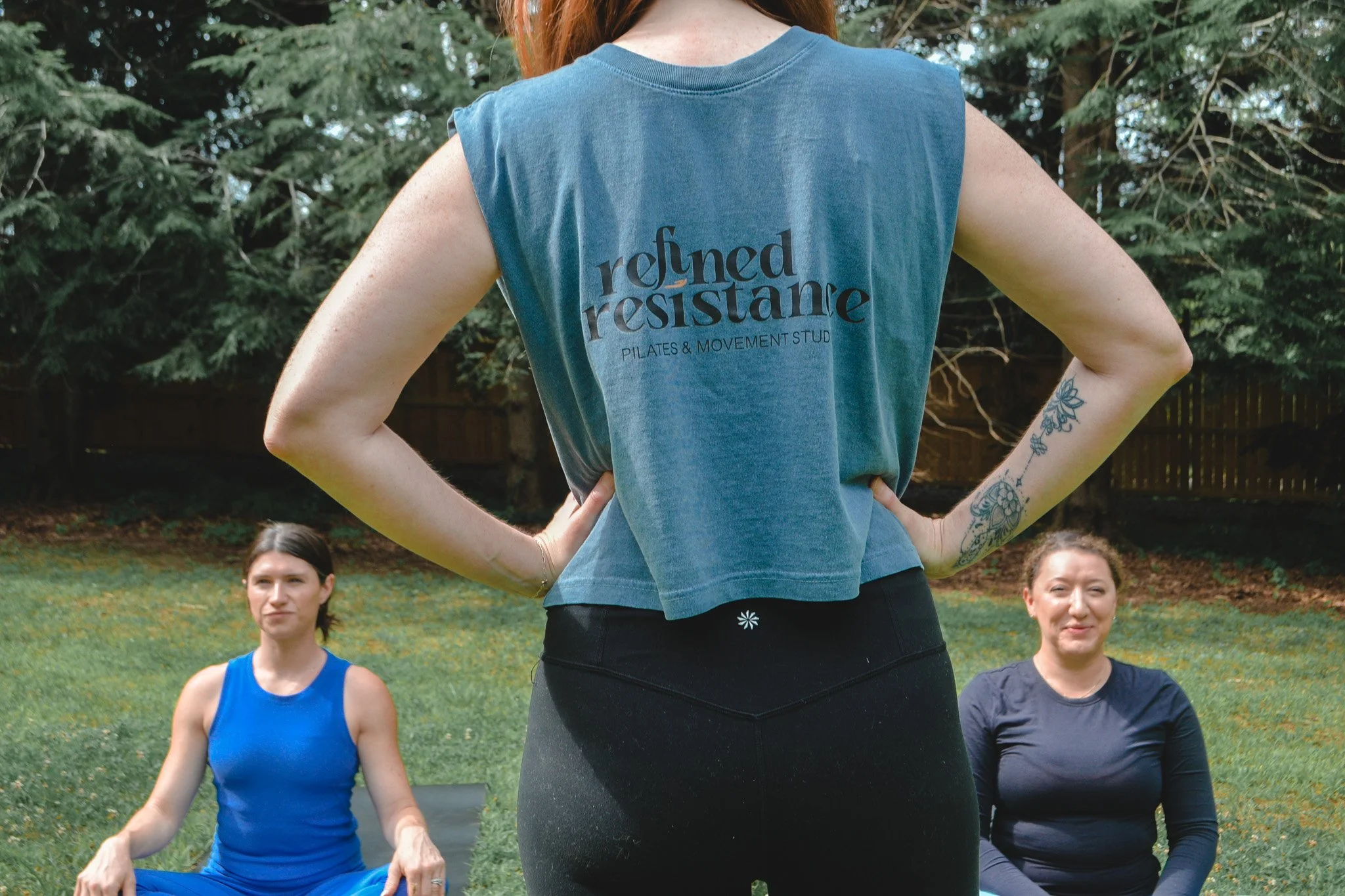 Women participating in an outdoor fitness class, sitting in a cross-legged position, facing their pilates instructor who is standing with her back to the camera wearing a shirt that says Refined Resistance Pilates and Movement Studio.