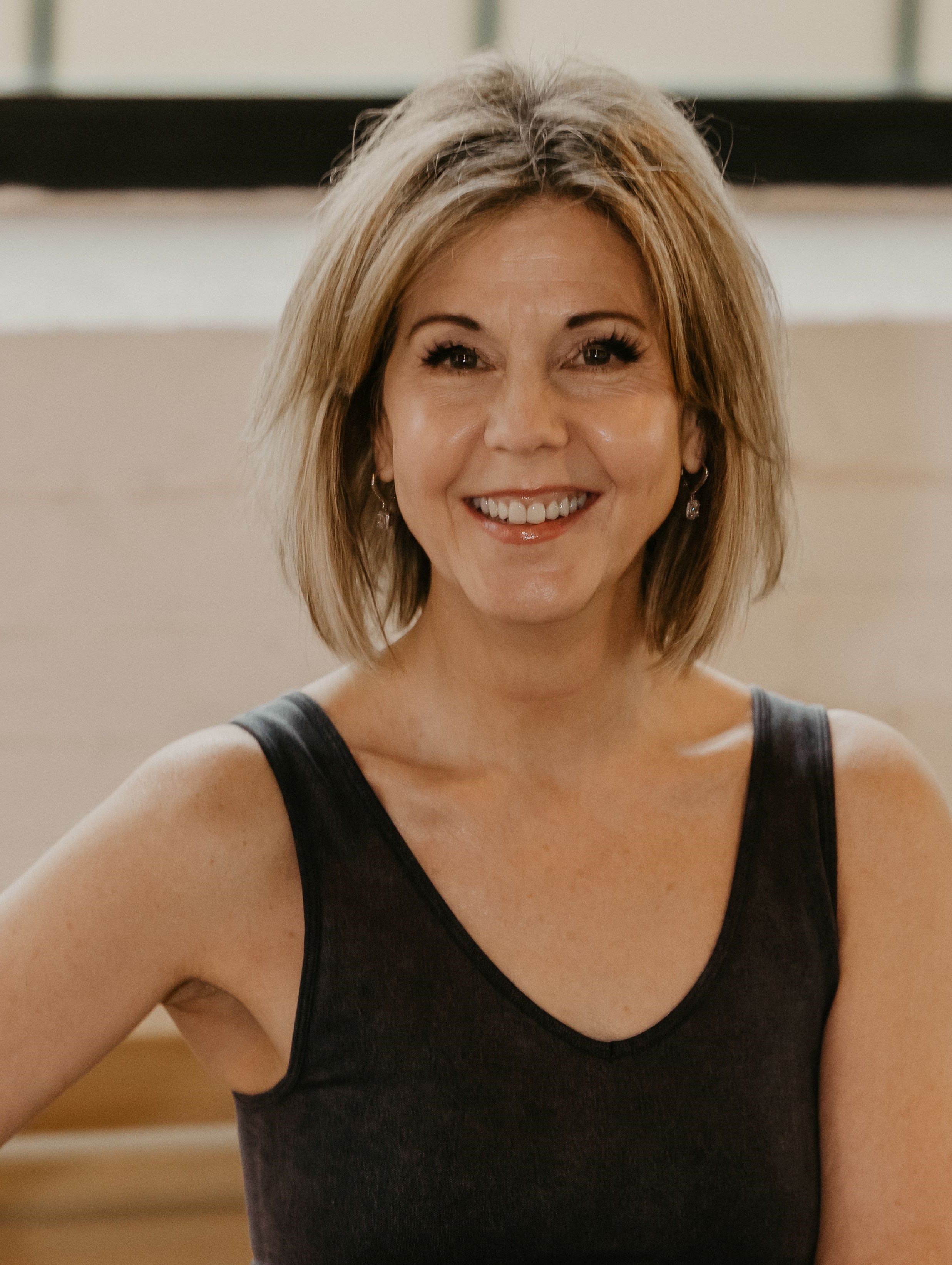 A woman who is a pilates instructor with blonde hair smiling, wearing a black sleeveless top, sitting indoors.