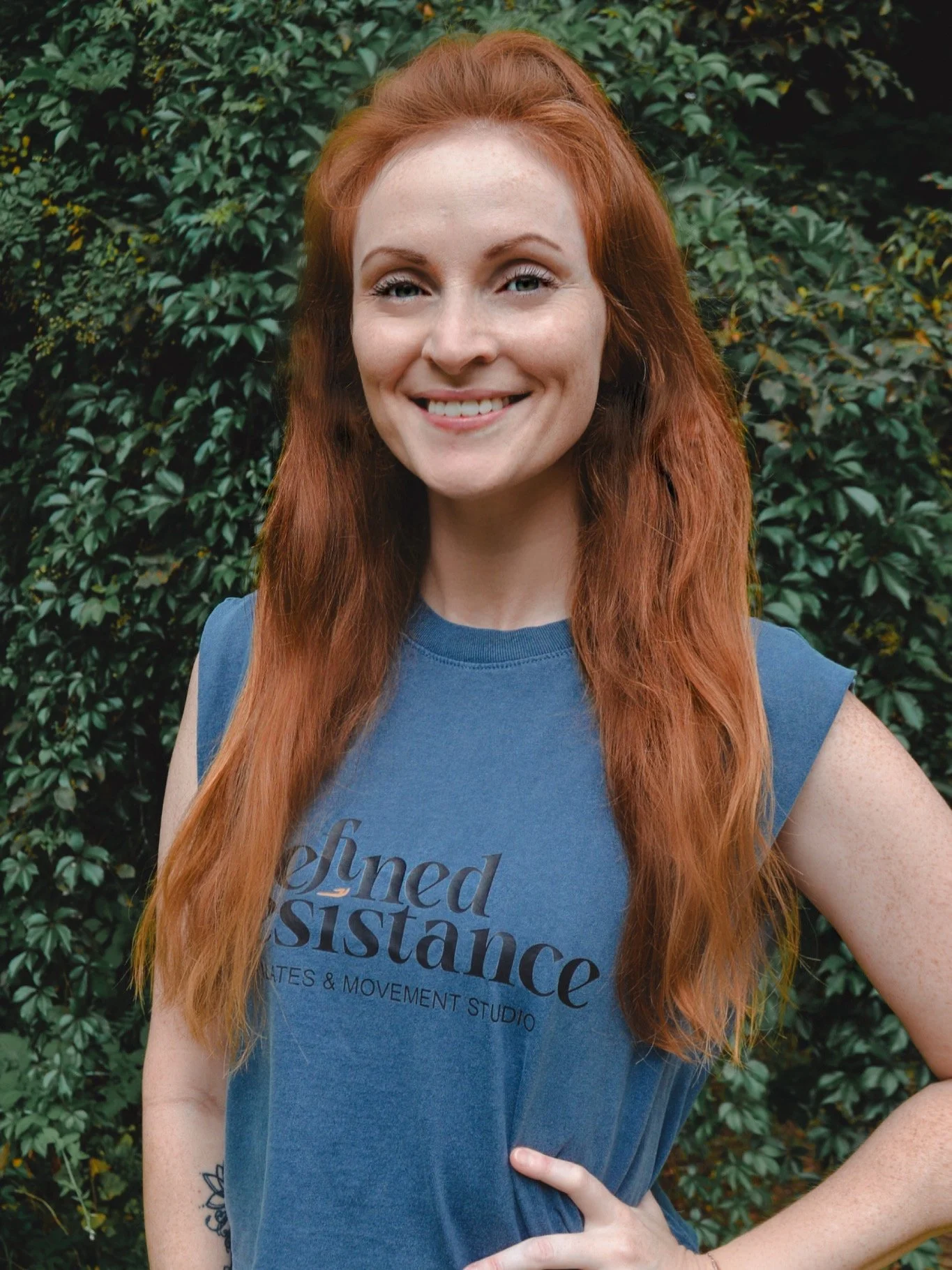 A woman who is a pilates instructor with long red hair and fair skin smiling outdoors with green bushes in the background, wearing a blue t-shirt.