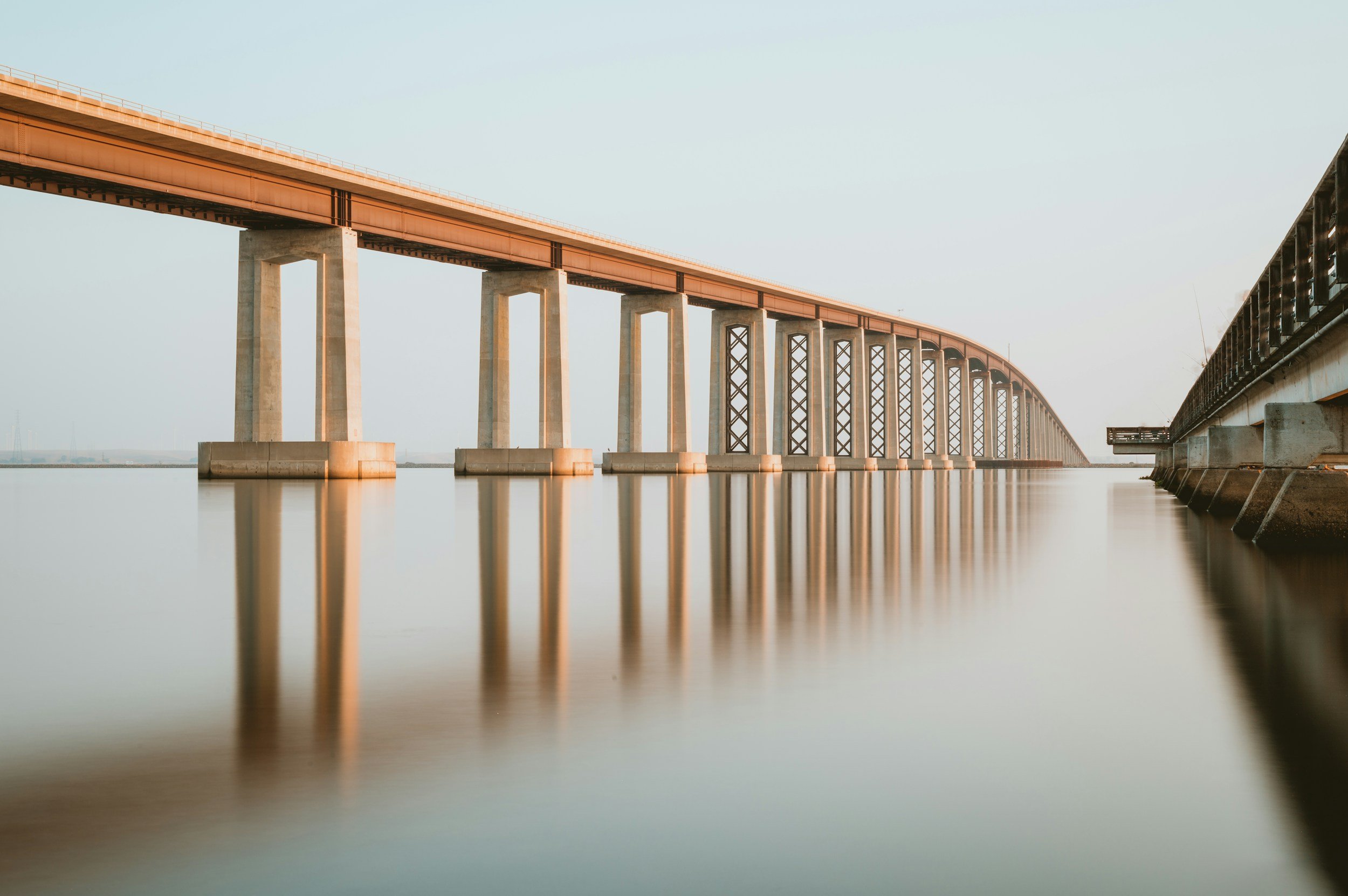 A long bridge with concrete pillars extending over a calm body of water, with a clear sky in the background.