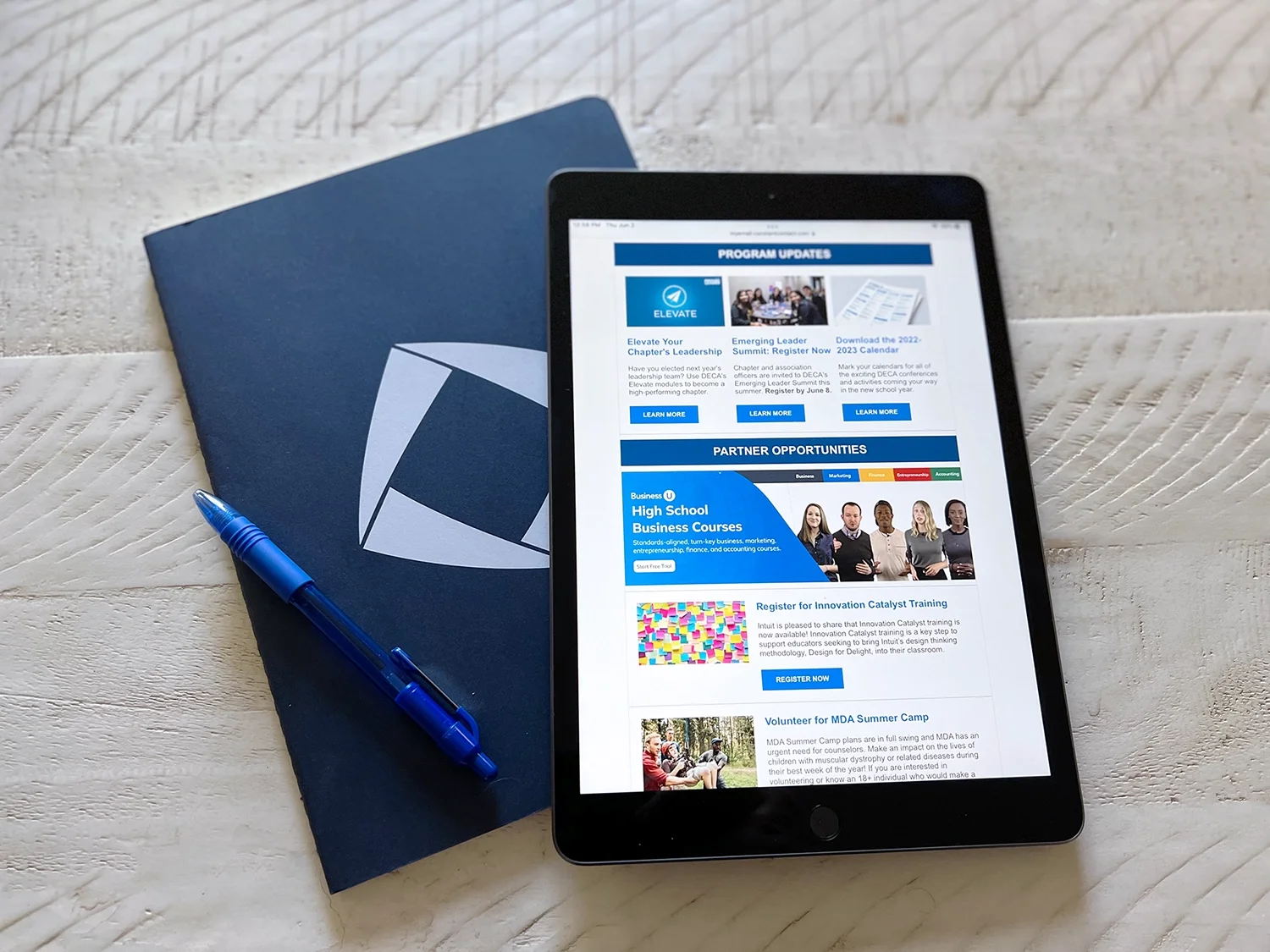Digital tablet displaying program updates and partner opportunities next to a blue notebook with a white geometric design, on a white textured wooden surface, with a blue pen resting on the notebook.