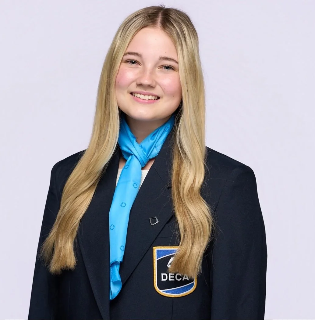 A young woman with long blonde hair smiling, wearing a black blazer with a DECA patch and a blue scarf.