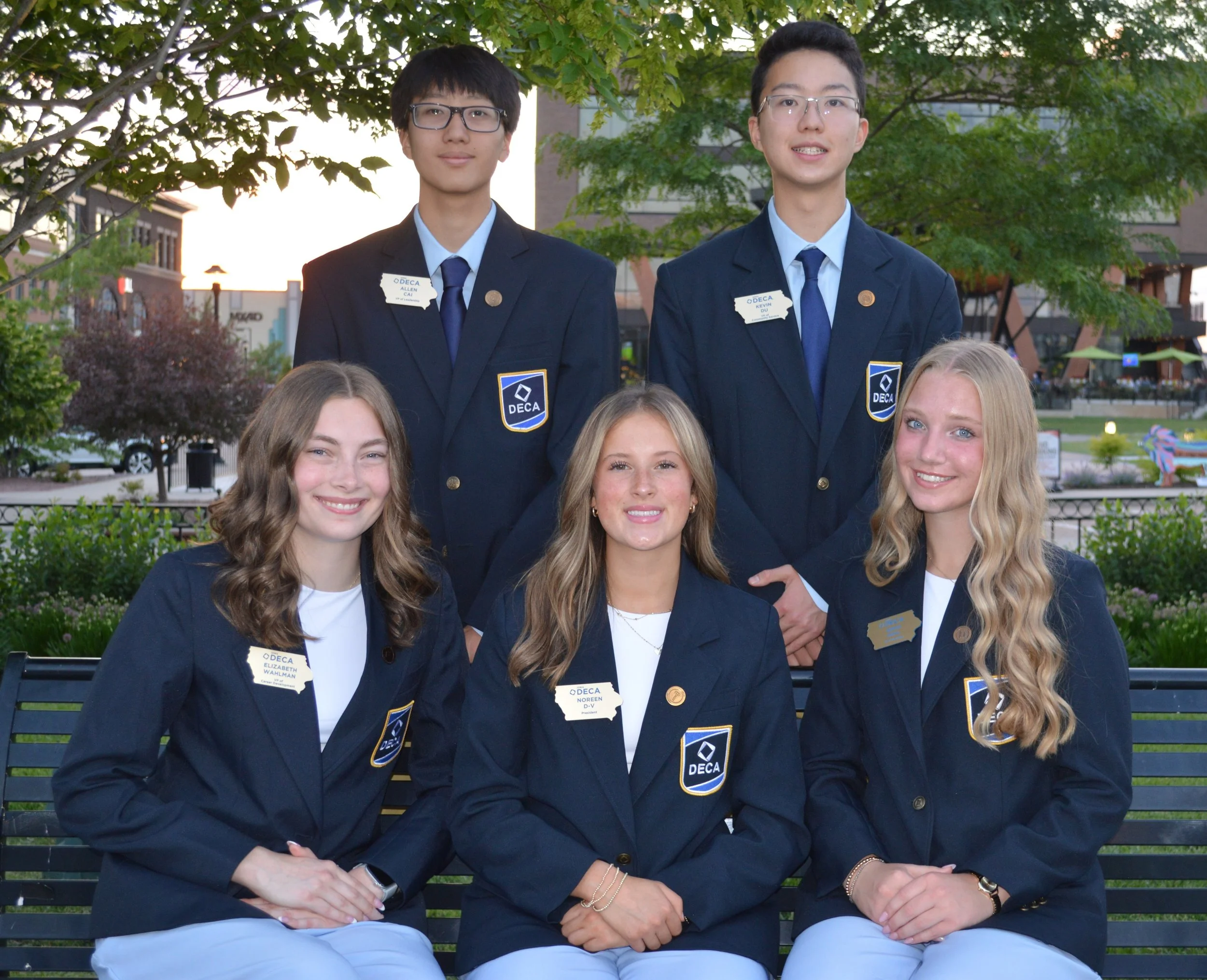 A group of young students in navy blazers with DECA badges, sitting and standing outdoors with trees and a building in the background.