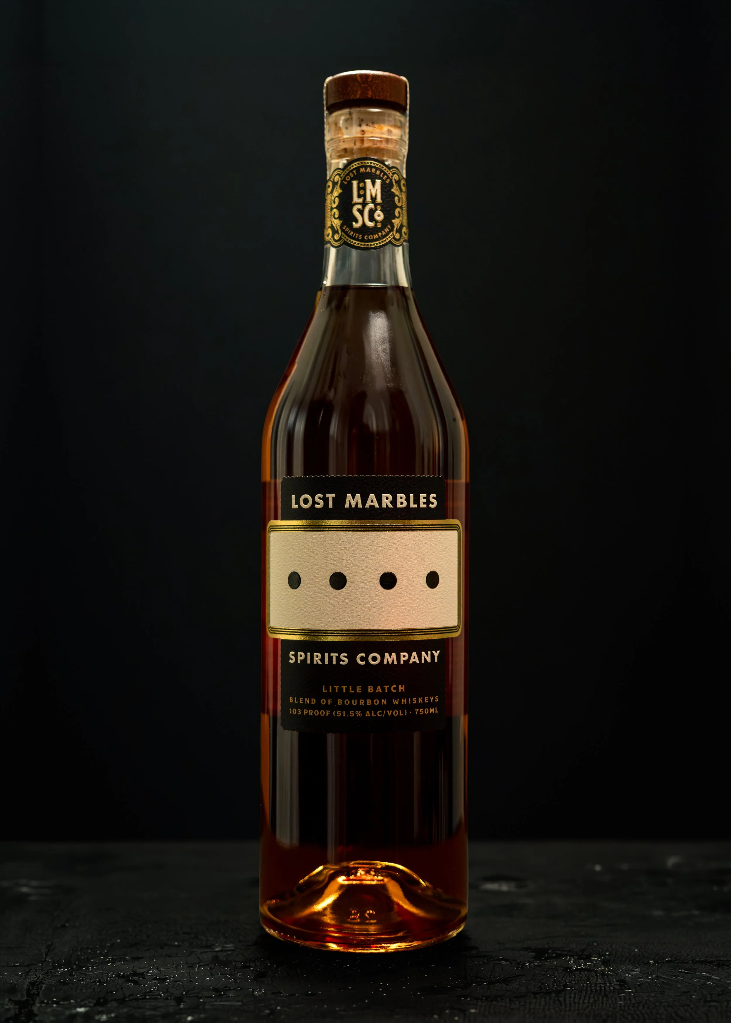 A tall glass bottle of Lost Marbles spirits with a cork top, a black and gold label, and a beige paper patch with four black dots, set against a dark background and placed on a textured dark surface.