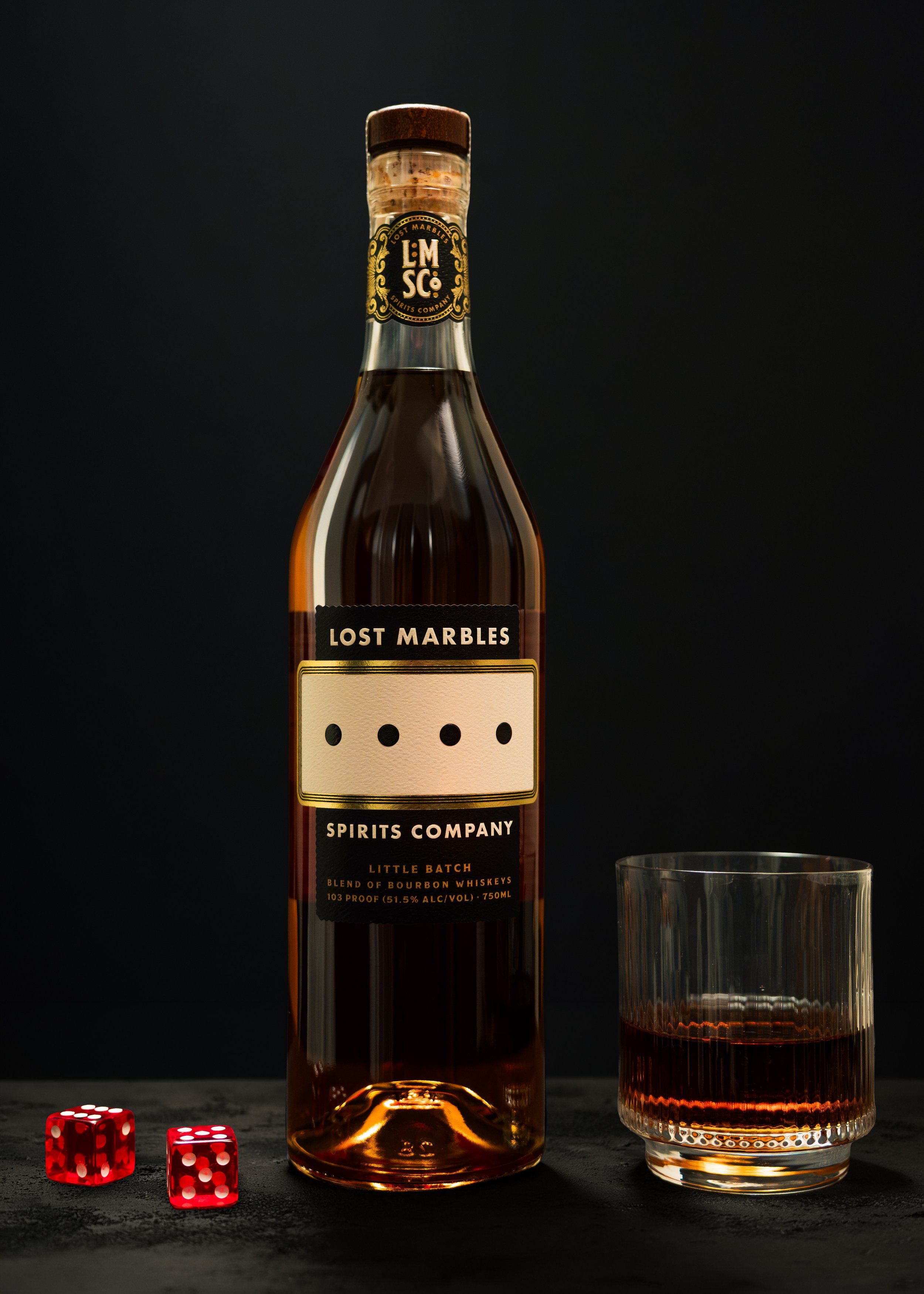 A bottle of Lost Marbles bourbon whiskey next to a glass filled with the same liquor, with two red dice showing the numbers six and five, on a dark textured surface against a black background.