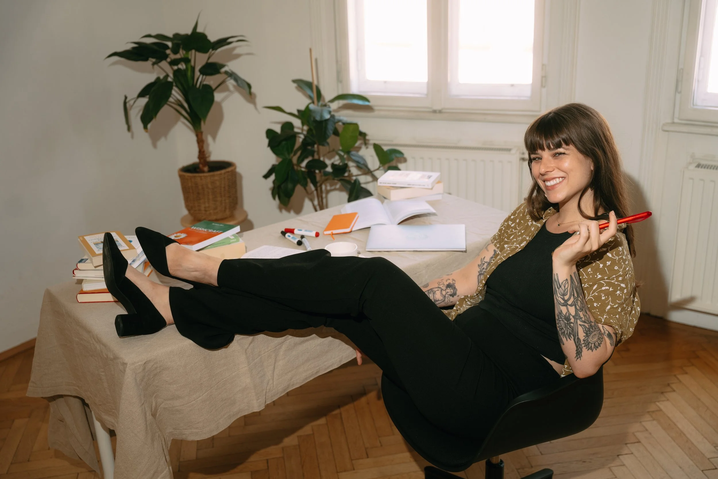A woman with tattoos on her arms and long brown hair, smiling and sitting with her legs stretched out on a table covered with books, papers, and pens in a room with natural light, a window, and potted plants.