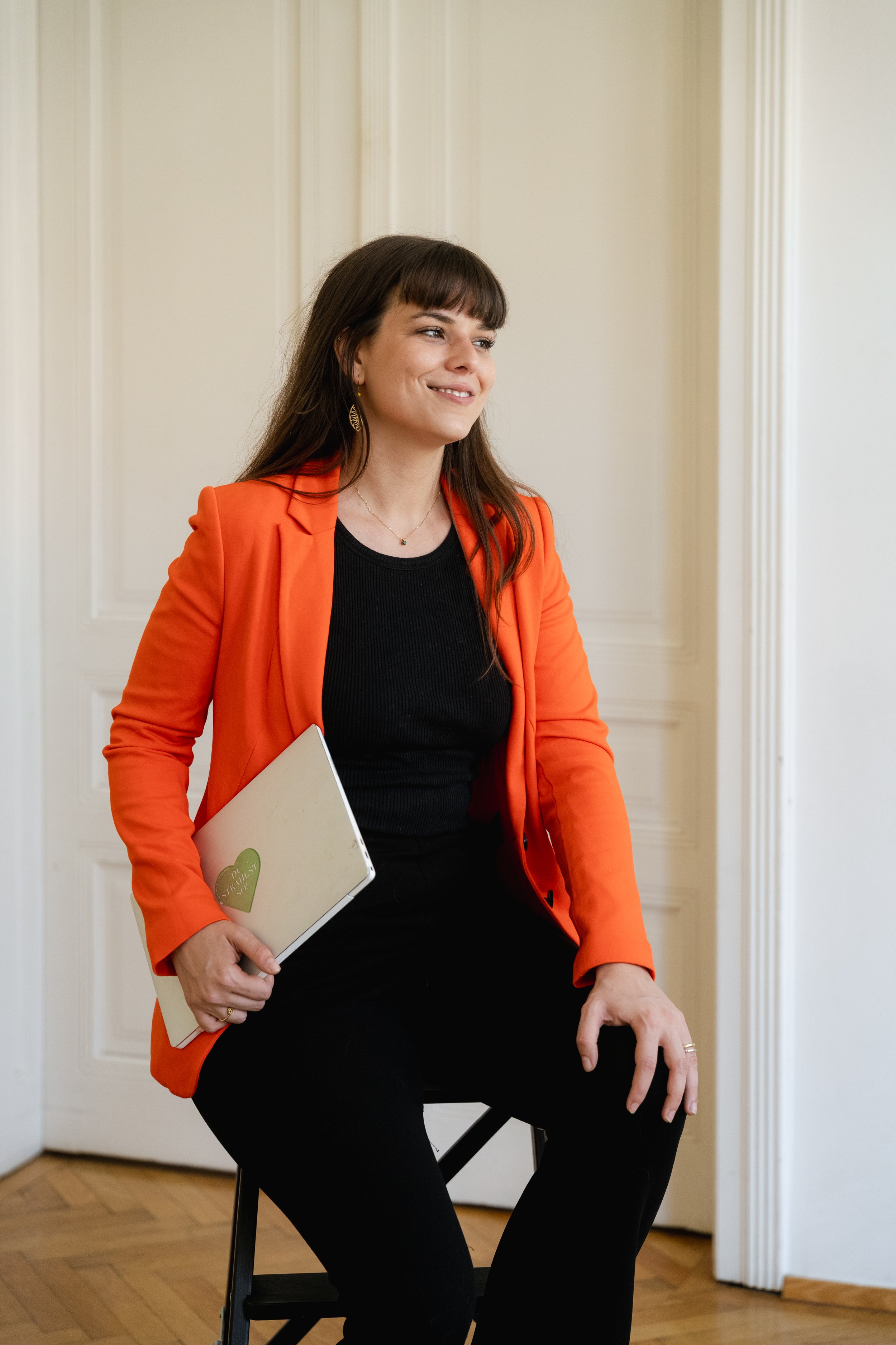 A woman with brown hair and bangs, wearing a black top and an orange blazer, sits on a black chair holding a closed laptop with a sticker that says 'Spread Love' and a green heart. She is smiling and looking to her left, in a room with wooden flooring and white paneled walls.