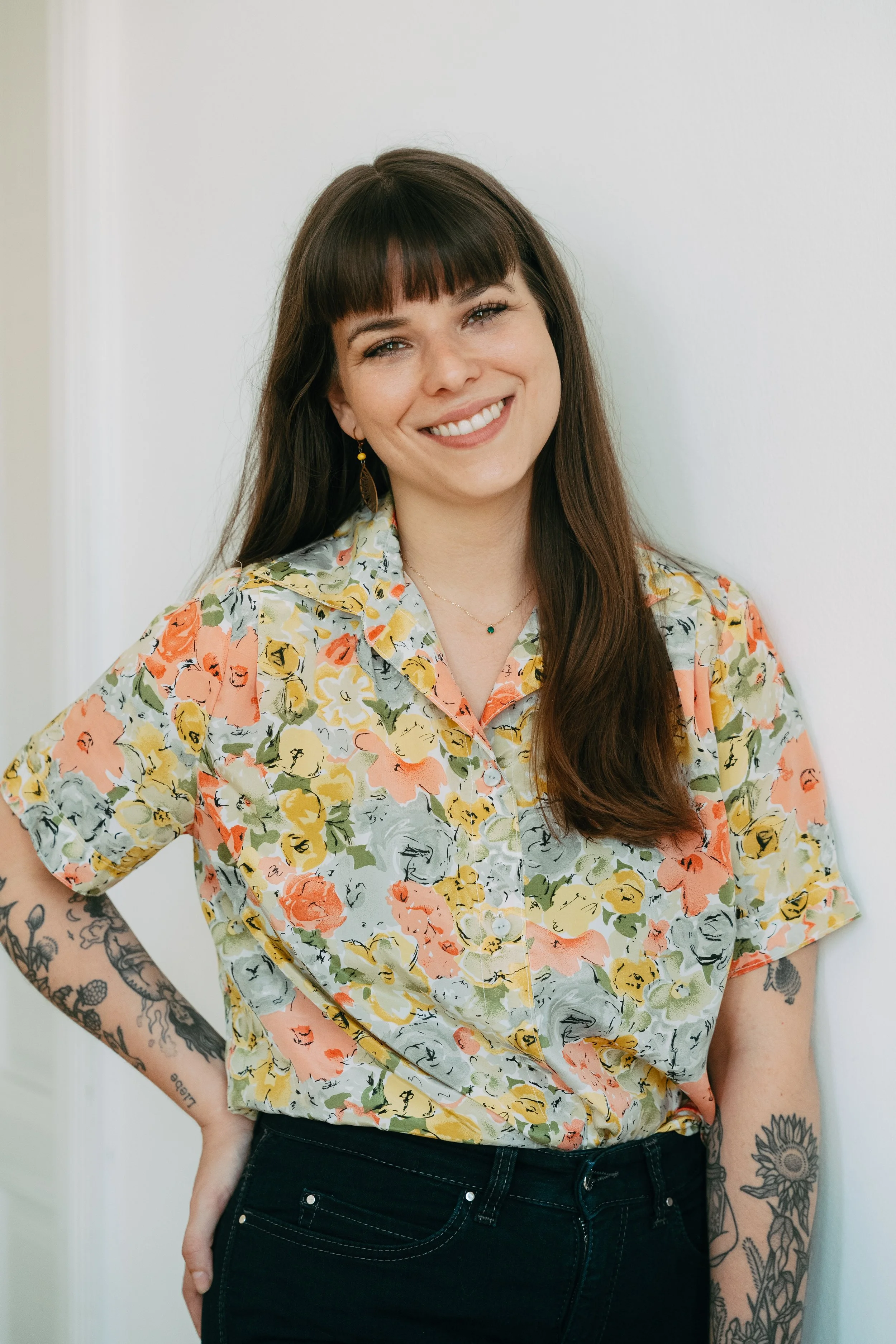 A woman with long brown hair, wearing a floral short-sleeve button-up shirt and black jeans, smiling and standing against a white wall, with tattoos on her left arm.