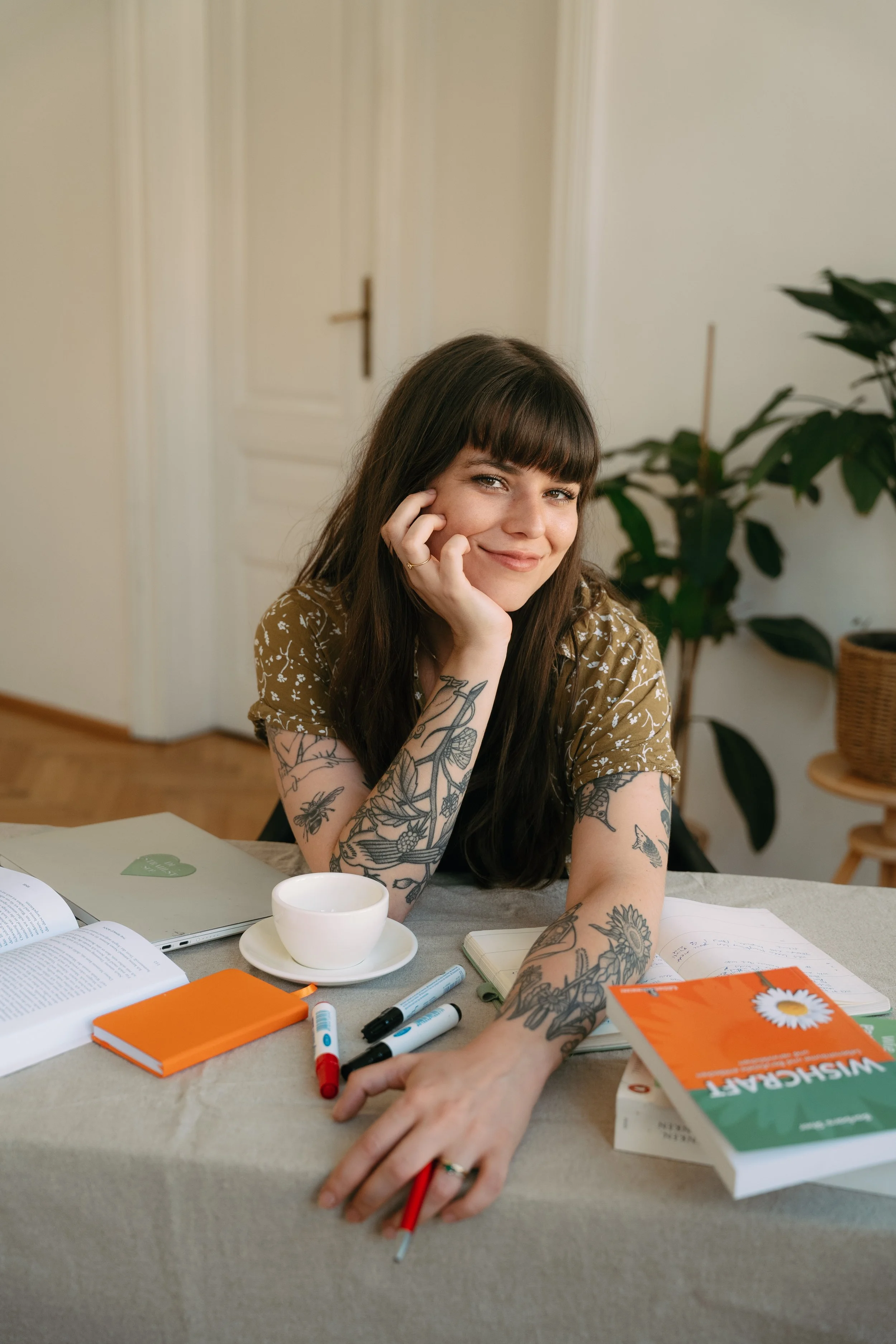 A young woman with long dark hair and tattoos on her arms, smiling and leaning her face on her hand, sitting at a table with notebooks, pens, a book titled "Wissenschaft", a cup, and various writing supplies. The background includes a large plant and a closed door.