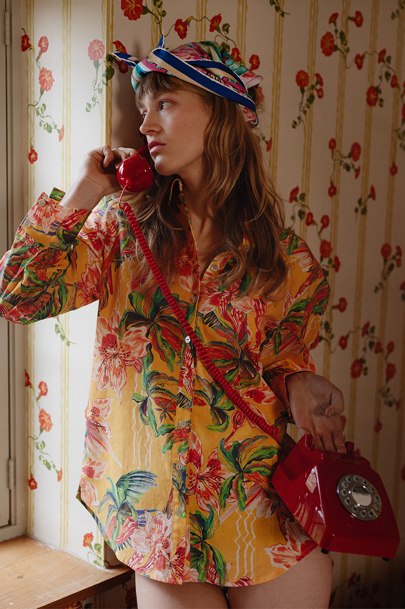 A woman with long brown hair wearing a colorful, floral-patterned shirt and a patterned headscarf, holding a red rotary phone to her ear while standing in a room with floral wallpaper.