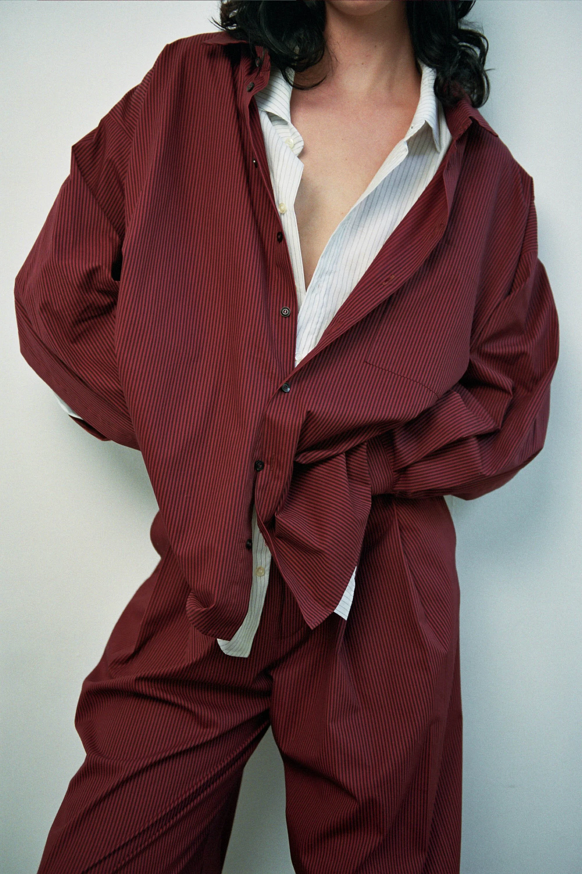 Close-up of a woman wearing a red pinstripe suit with a white dress shirt, with her arms crossed in front of her, against a plain light background.