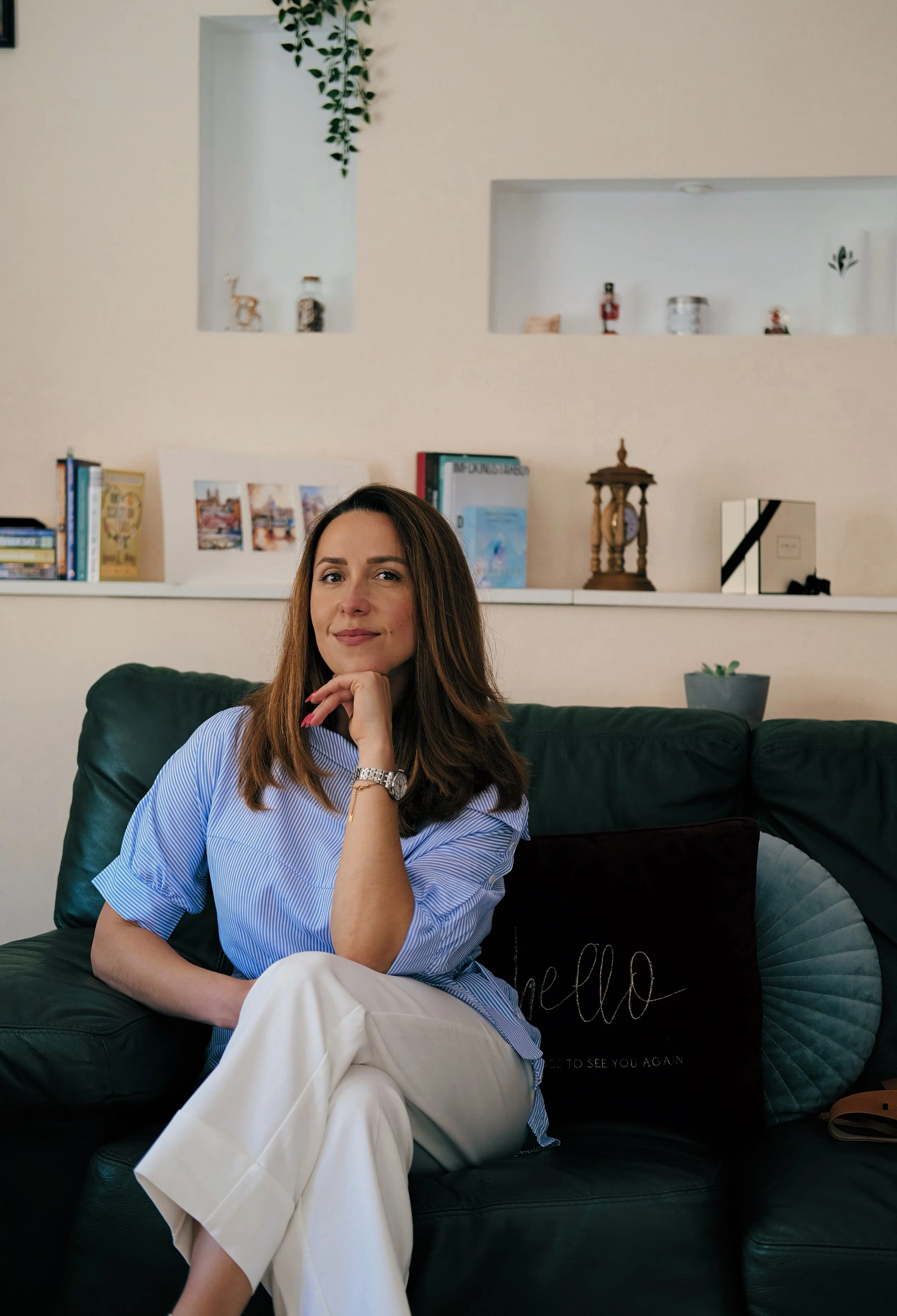 A woman with long brown hair sitting on a dark green leather sofa, wearing a light blue striped shirt and white pants, with her chin resting on her hand. Behind her is a white wall with books, framed pictures, a vintage clock, and decorative items displayed on a shelf. There is a gray pillow with the word 'hello' written on it next to her.