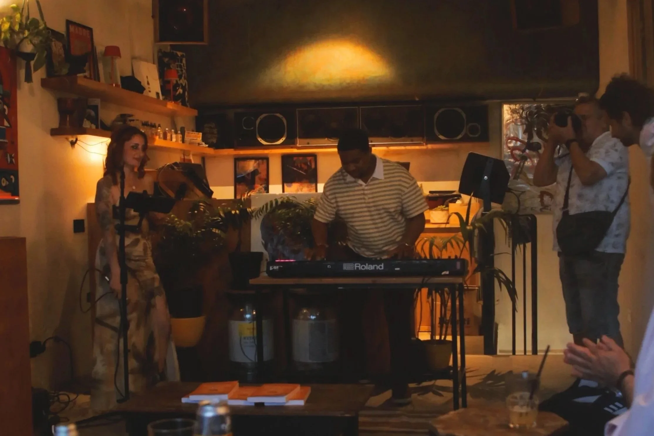 People in a dimly lit room, one woman and two men, with Grace WILDE singing, Gavin Rushing playing a keyboard and another filming or taking photos at RELENTLESS: Not Your Usual Book Launch