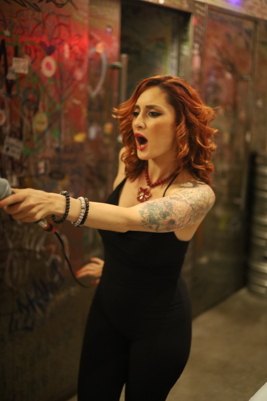Grace WILDE in a black dress, holding a microphone, in a colorful, graffiti-covered environment.