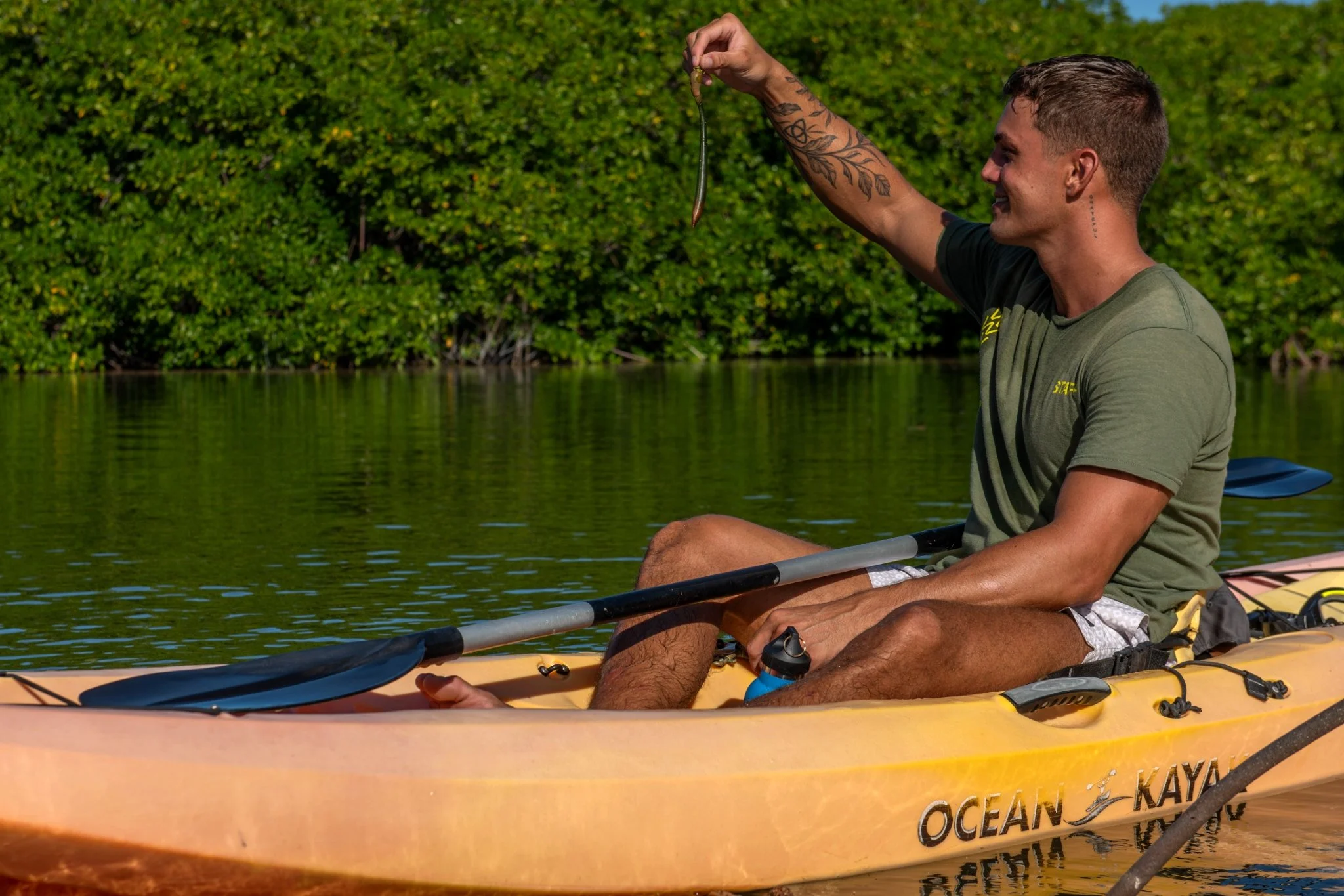 A man sitting in a kayak on a calm body of water, holding a small snake aloft in his right hand, smiling at it. The kayak has the words 'Ocean Kayak' on its side, and the background shows lush green trees.