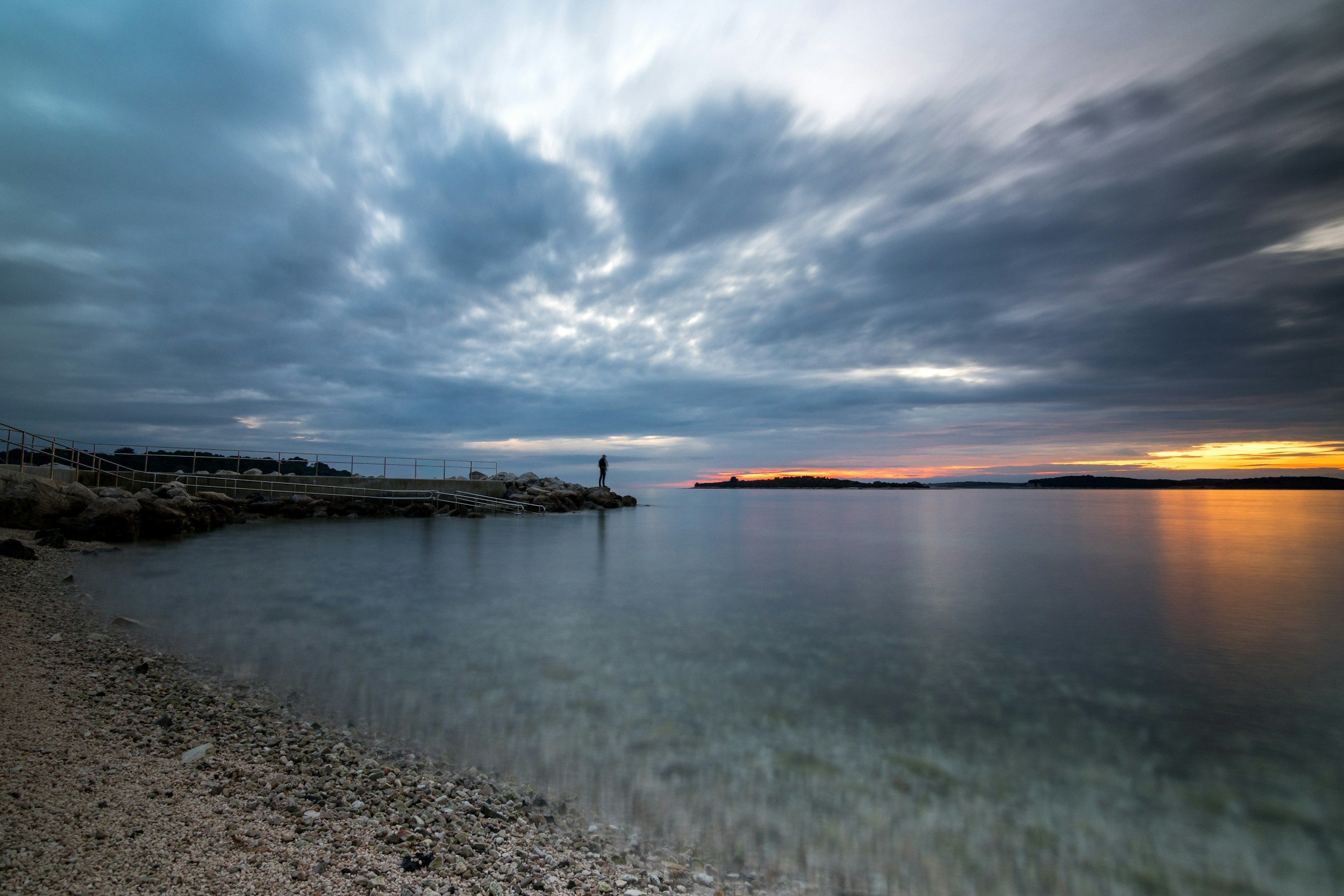 A person in the distance standing on land jutting into water looking out toward another piece of land across the water.
