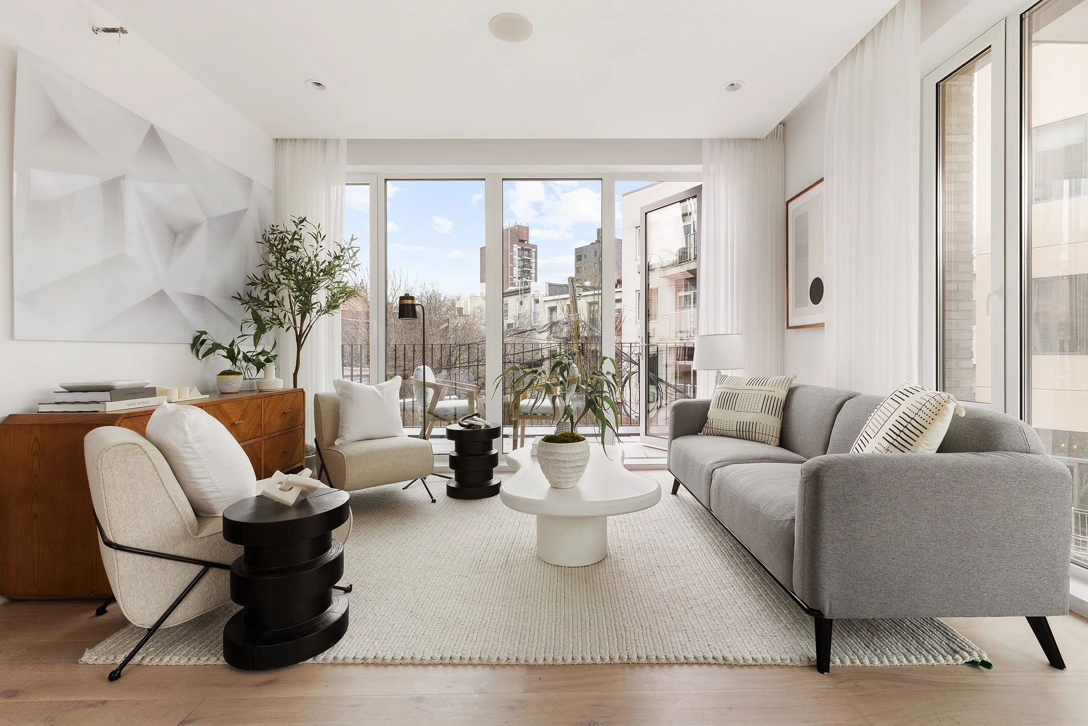 Modern living room with large windows, gray sofa, beige armchairs, white coffee table with a potted plant, black side tables, a wooden sideboard, artwork on white walls, and a balcony outside.