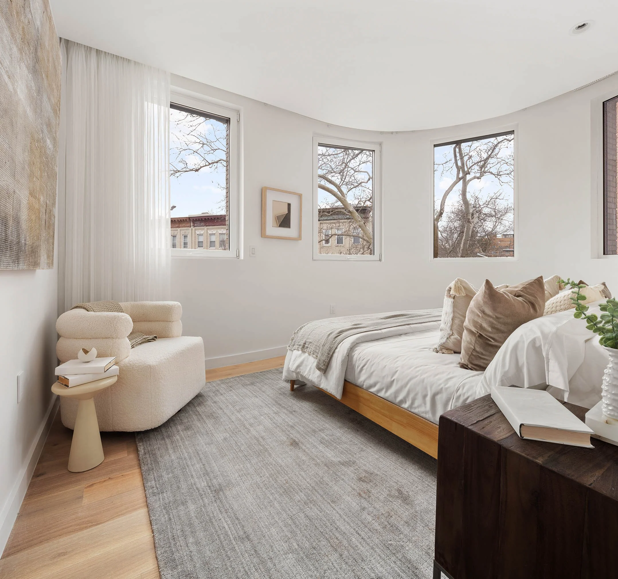 Bright bedroom with white walls, wooden flooring, and large windows showing a tree outside. Features include a bed with beige and white bedding, a cream armchair, a small side table with books, and a dark wooden nightstand with books and a small plan