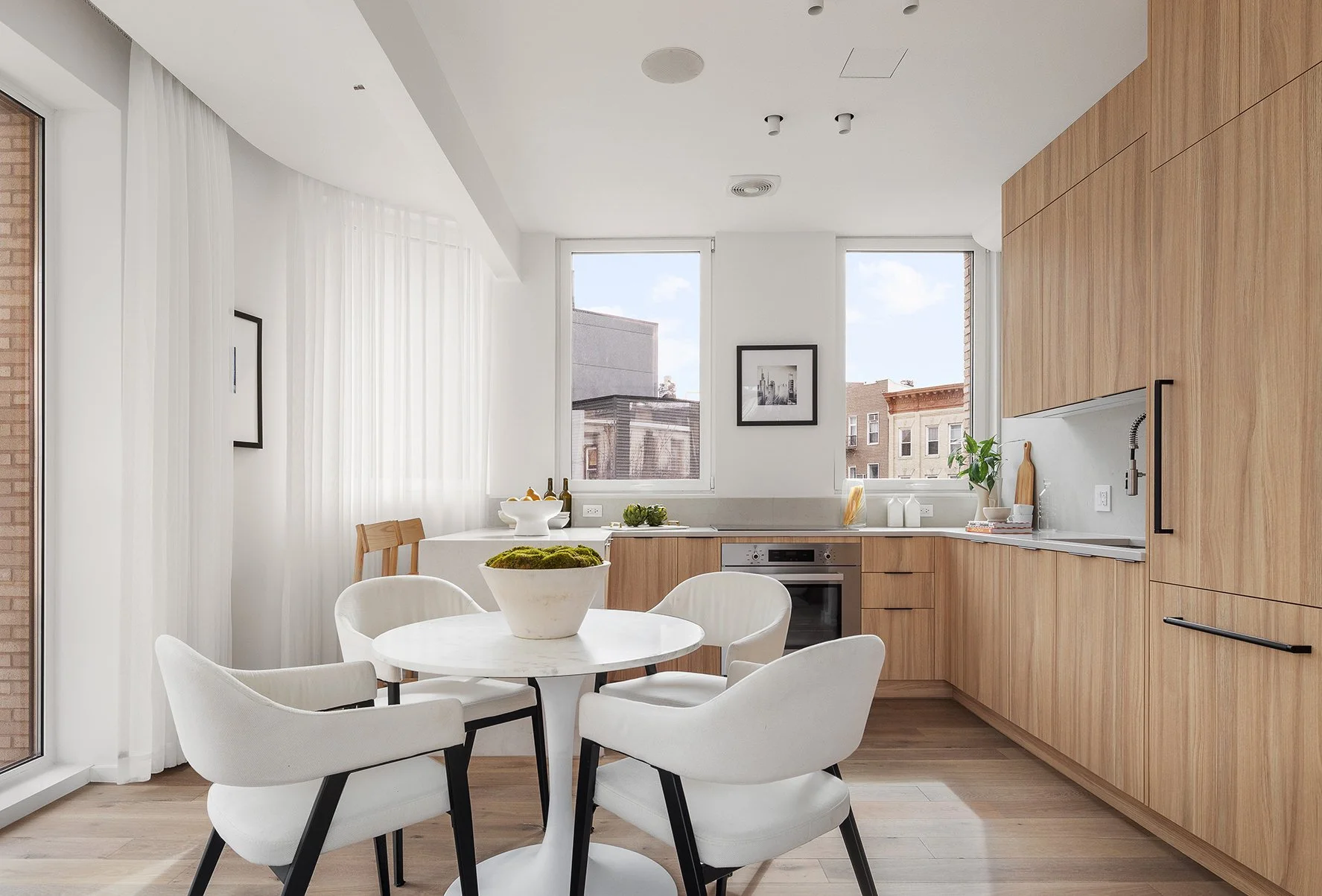 Modern kitchen with white walls, wooden cabinets, a round dining table with four white chairs, and large windows letting in natural light.