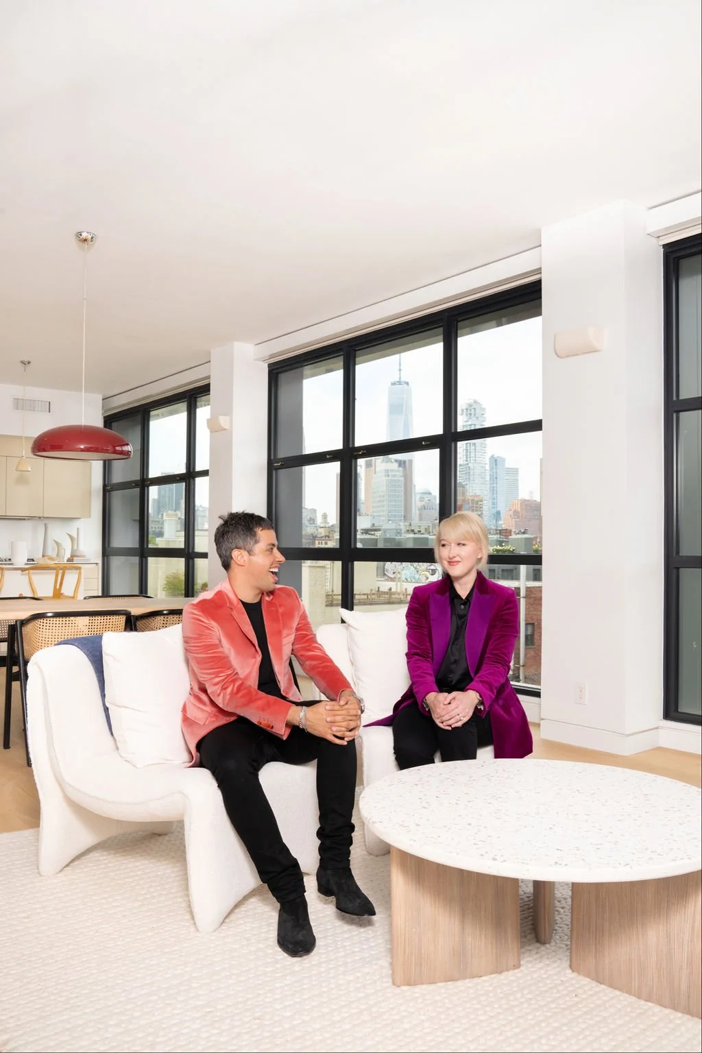Two women sitting on a white sofa in a modern urban apartment, having a conversation with a city skyline visible through large windows behind them.