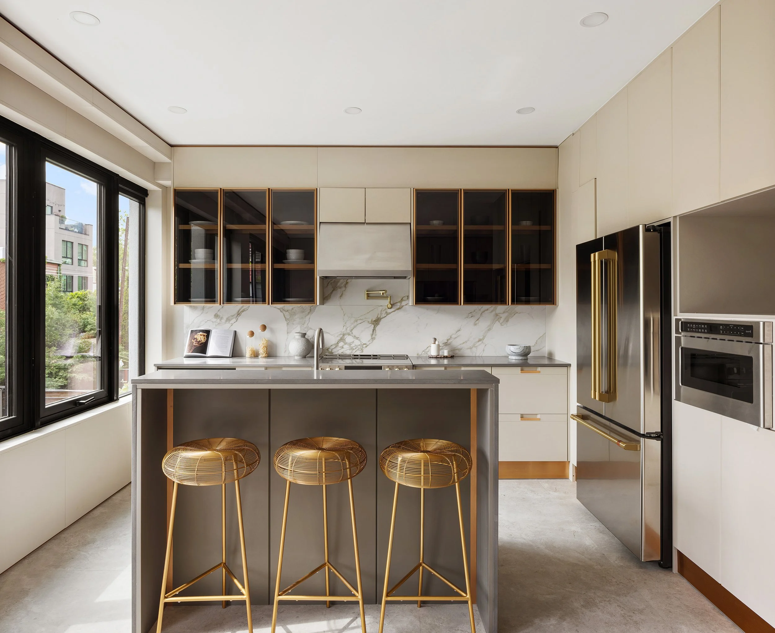 Modern kitchen with white marble backsplash, black window frames, gold accents on cabinetry and appliances, a marble island, and three gold wire stools