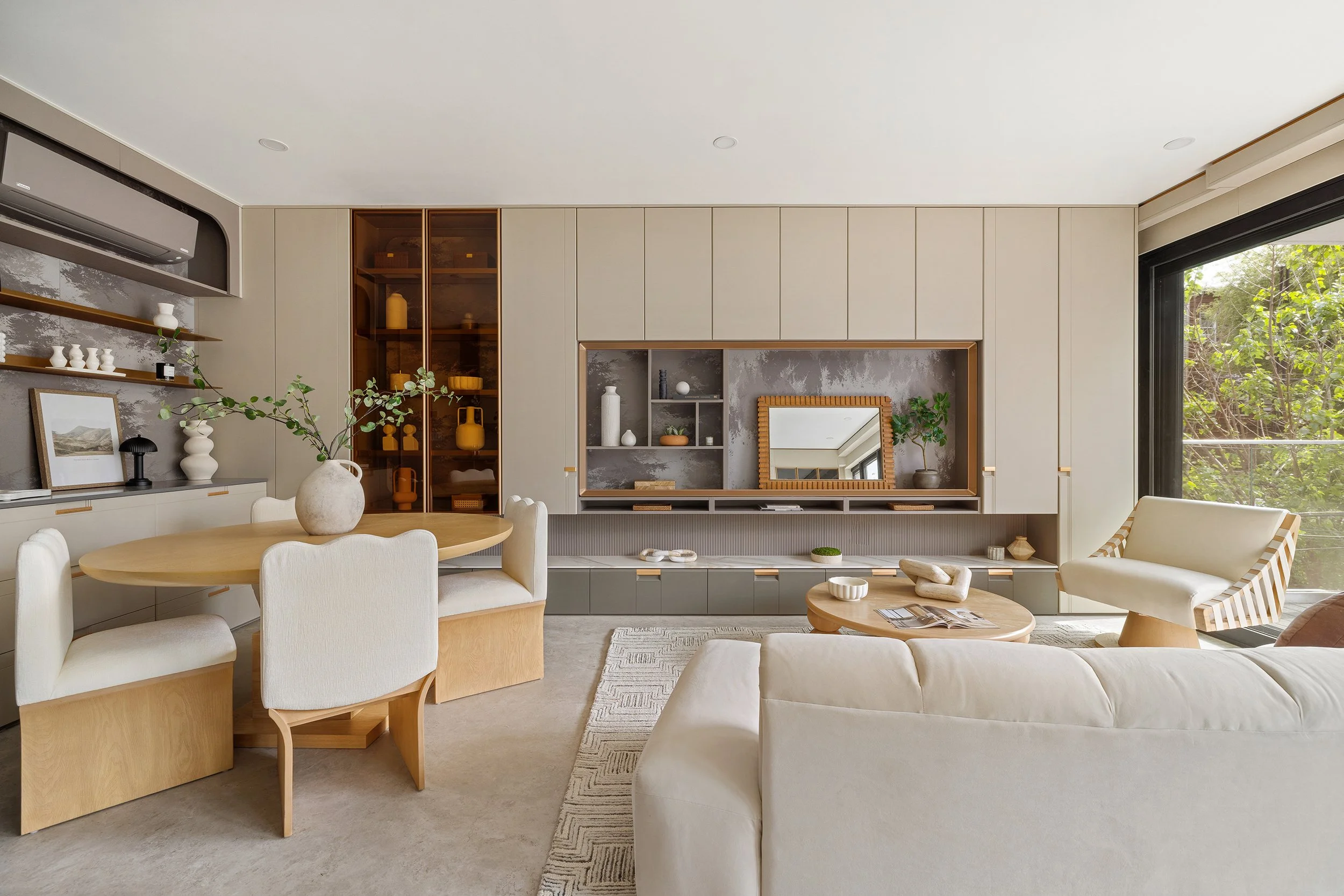 Modern living room with beige walls, built-in shelving, a large window with greenery outside, white sofa, wooden dining table with chairs, decorative vases, artwork, and plants.
