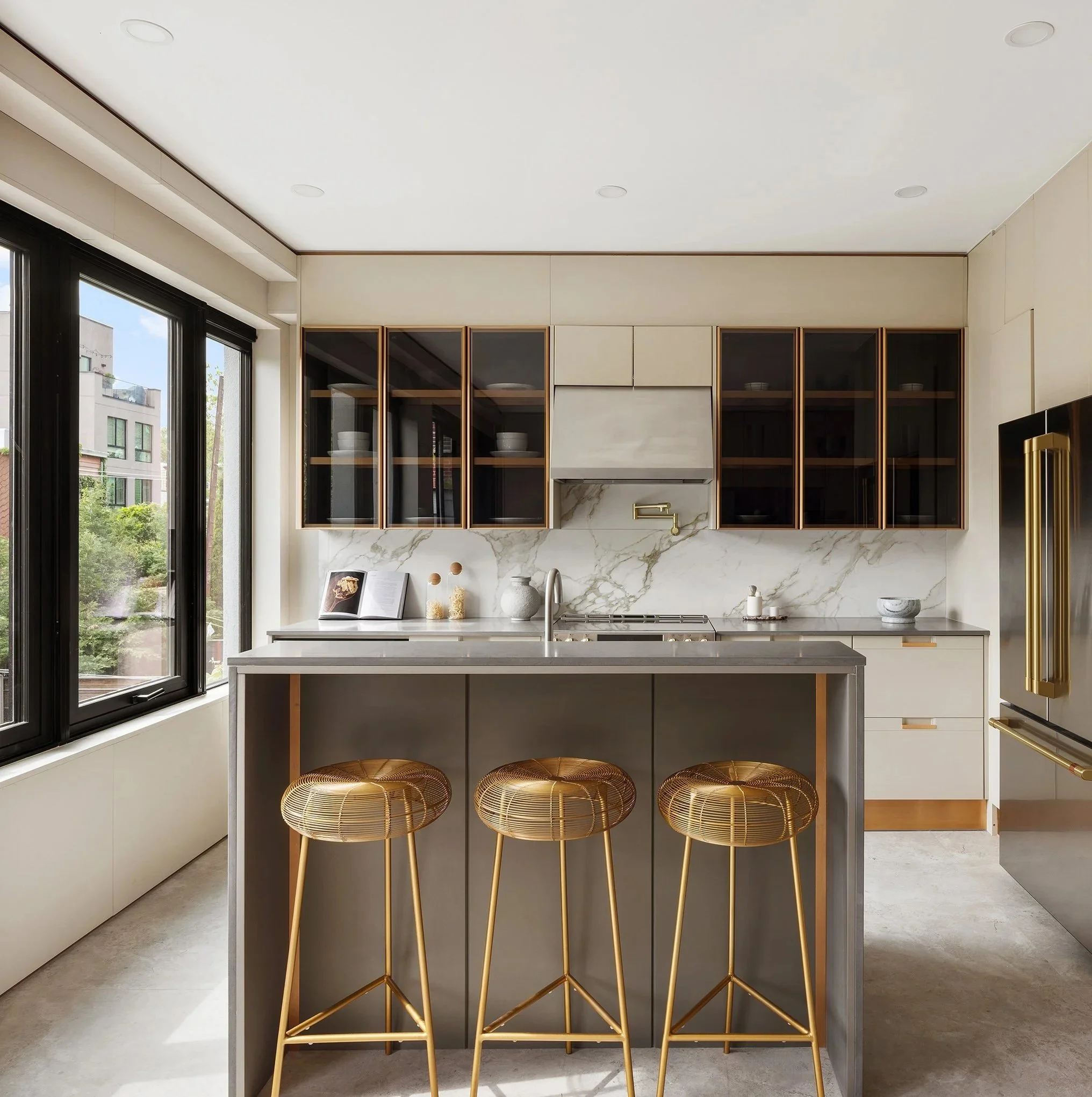Modern kitchen with beige cabinets, a marble backsplash, gold bar stools, and large windows showing trees and buildings outside.