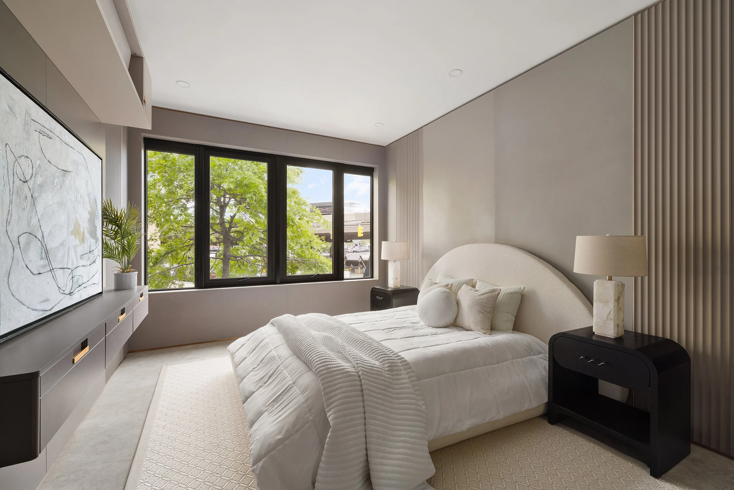 A modern bedroom with a large window showing green trees outside. The room has a white bed with pillows, a nightstand with a lamp, a black speaker, and abstract artwork on a console. The walls are decorated with textured panels.