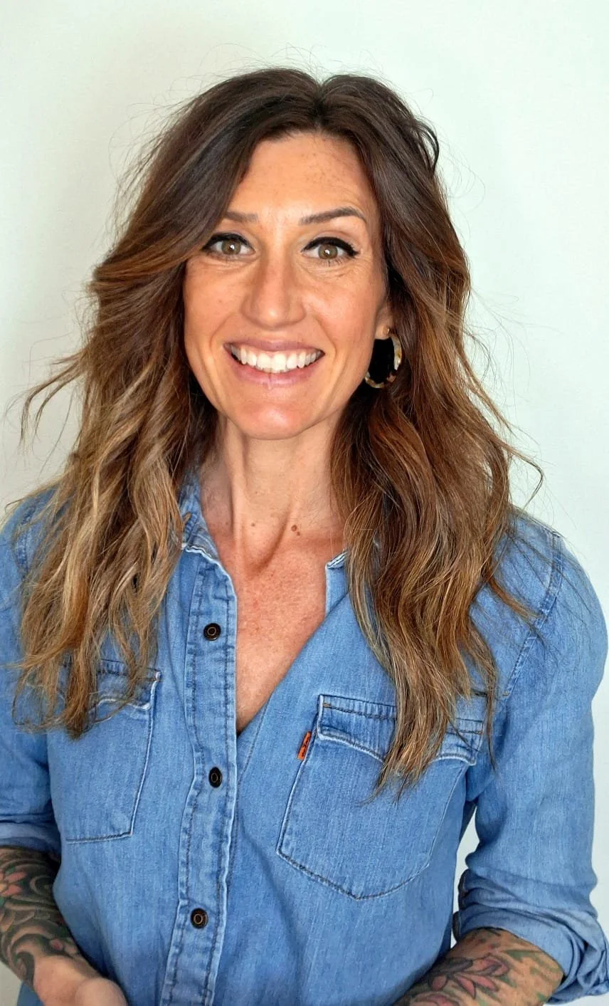A smiling woman with wavy brown hair, wearing a denim shirt and a hoop earring, standing against a plain white wall.