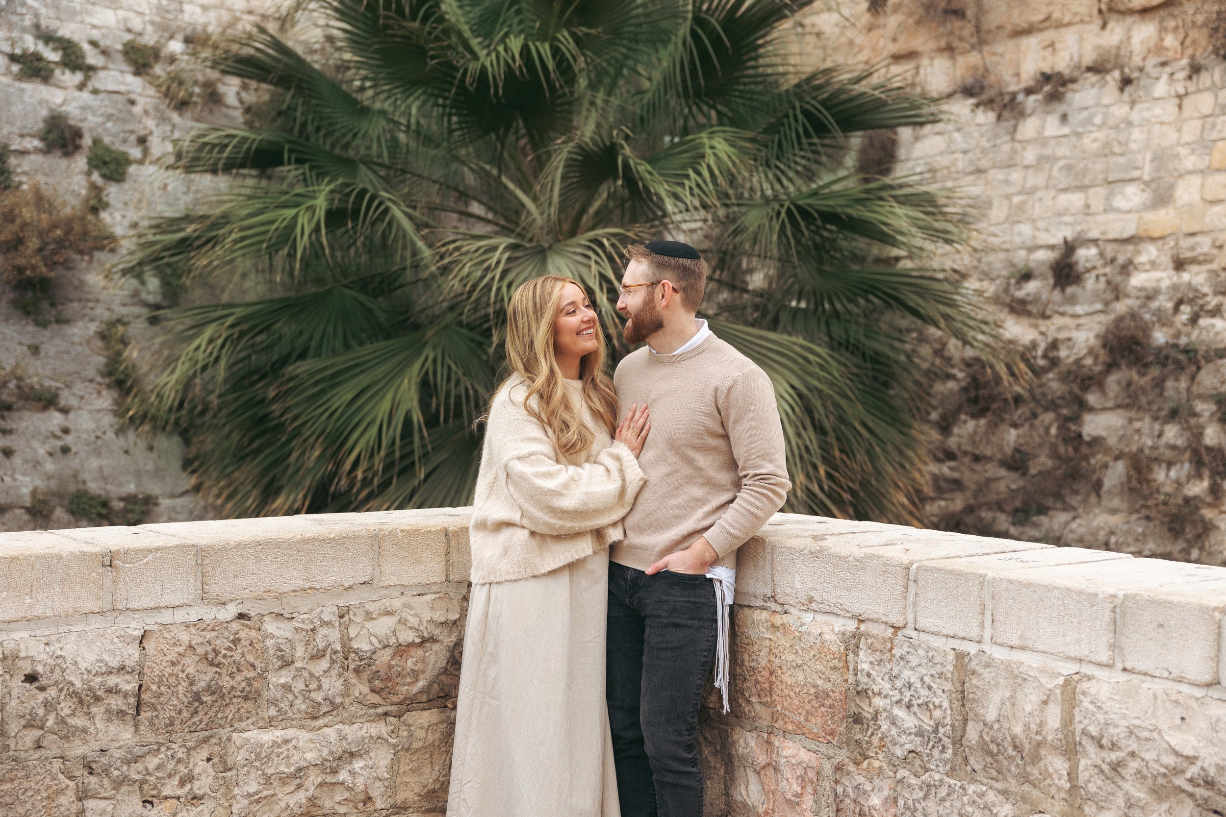 A couple stands close to each other in front of a stone wall and large green palm leaves. The woman has long blonde hair and is wearing a light-colored sweater and skirt, and the man has short hair, a beard, glasses, and is wearing a beige sweater and dark pants. They are smiling and looking at each other in a romantic moment.