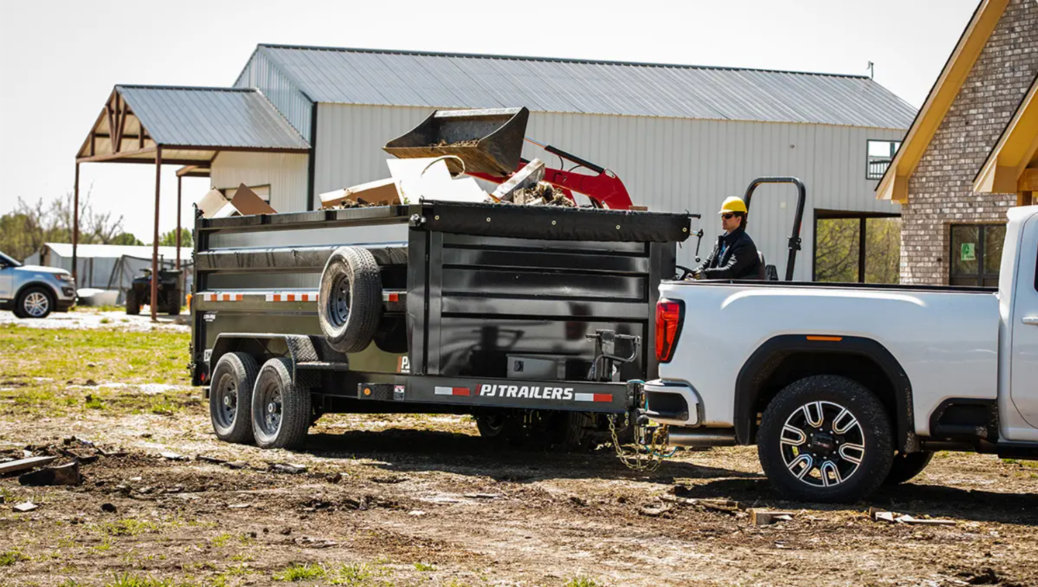 A construction worker in a yellow helmet operating a white pickup truck pulling a trailer filled with debris.