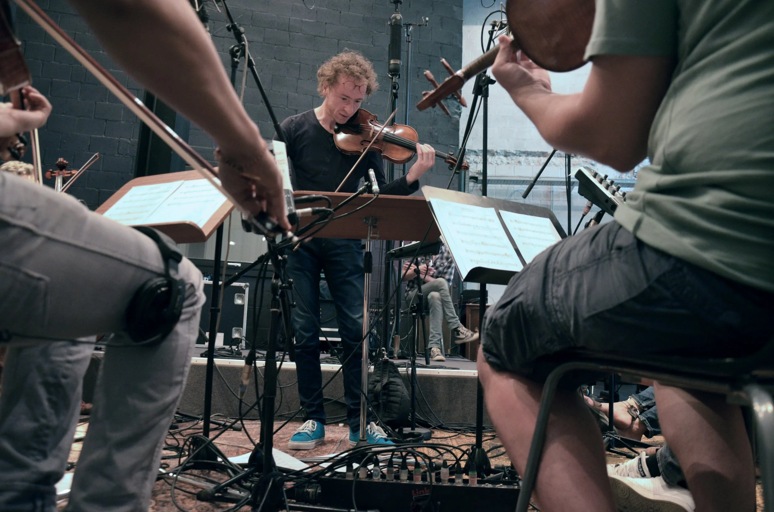 A musician with glasses and curly hair playing the violin during a recording session, surrounded by microphones and musical equipment, with other musicians and music stands visible in the background.