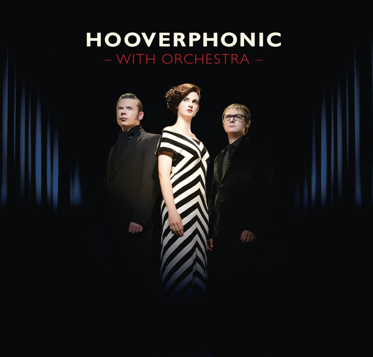 Promotional poster for 'Hoverphonic with Orchestra' featuring three people standing on stage, two men and one woman, with a dark background and blue curtains on the sides.