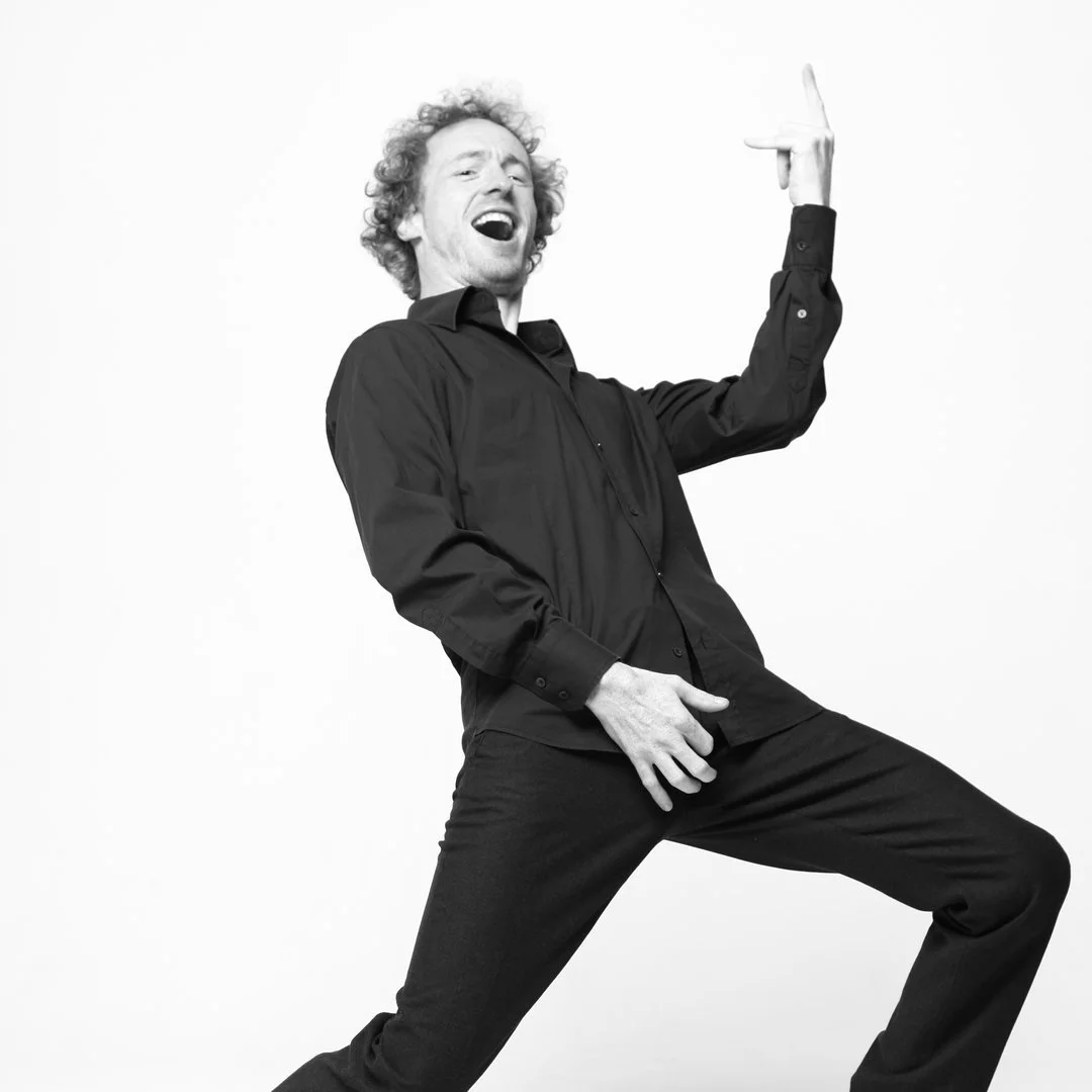 A man with curly hair dressed in black, posing with a lively expression and playing air guitar.