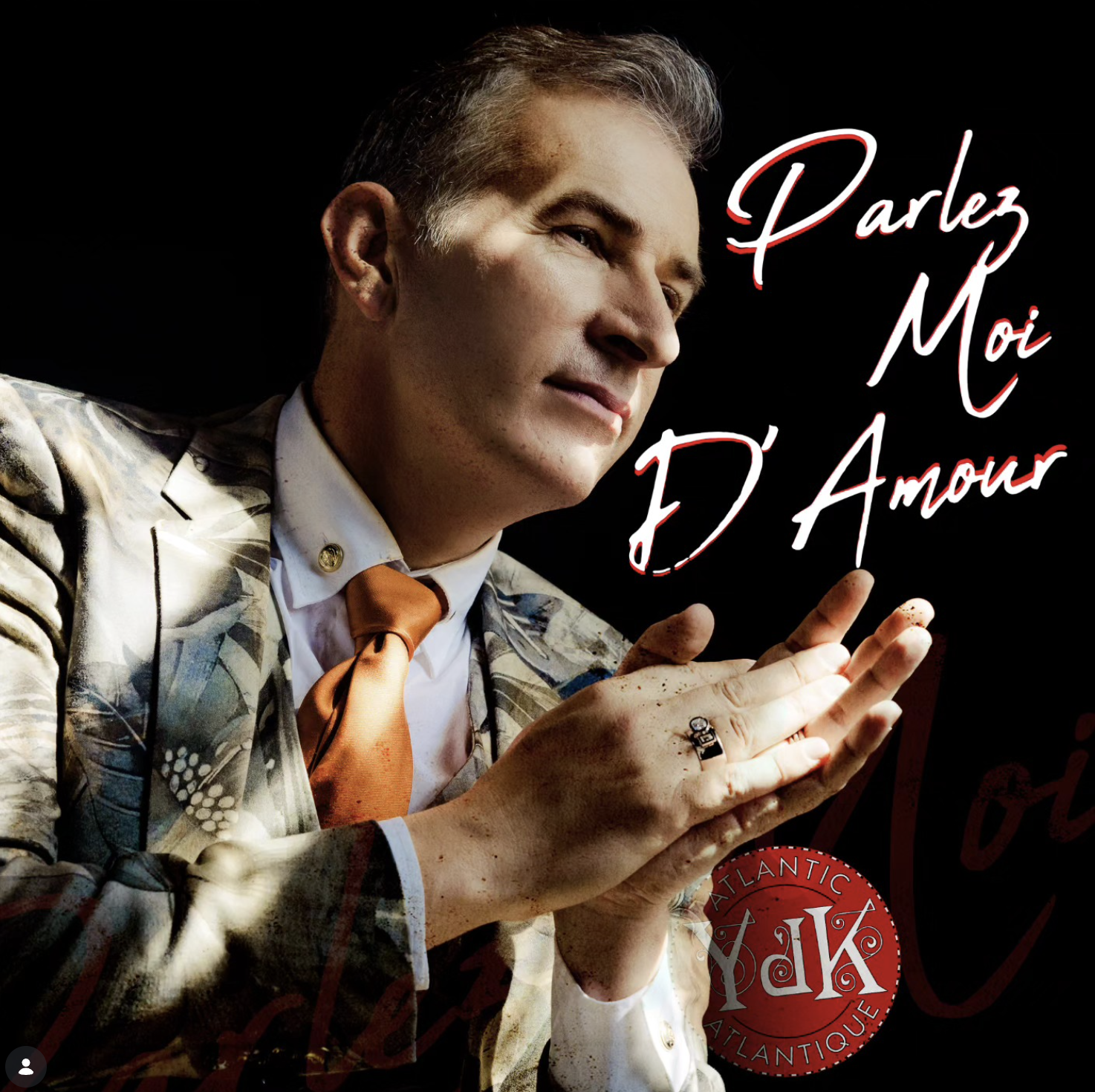 A man in a patterned blazer, white shirt, and orange tie has his hands together and looks thoughtfully. The image includes the text "Parlez Moi D'amour" in stylized red and white script.
