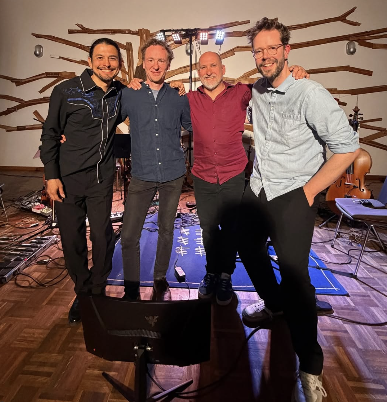 Four men standing together and smiling in a music studio with guitars and music equipment in the background.