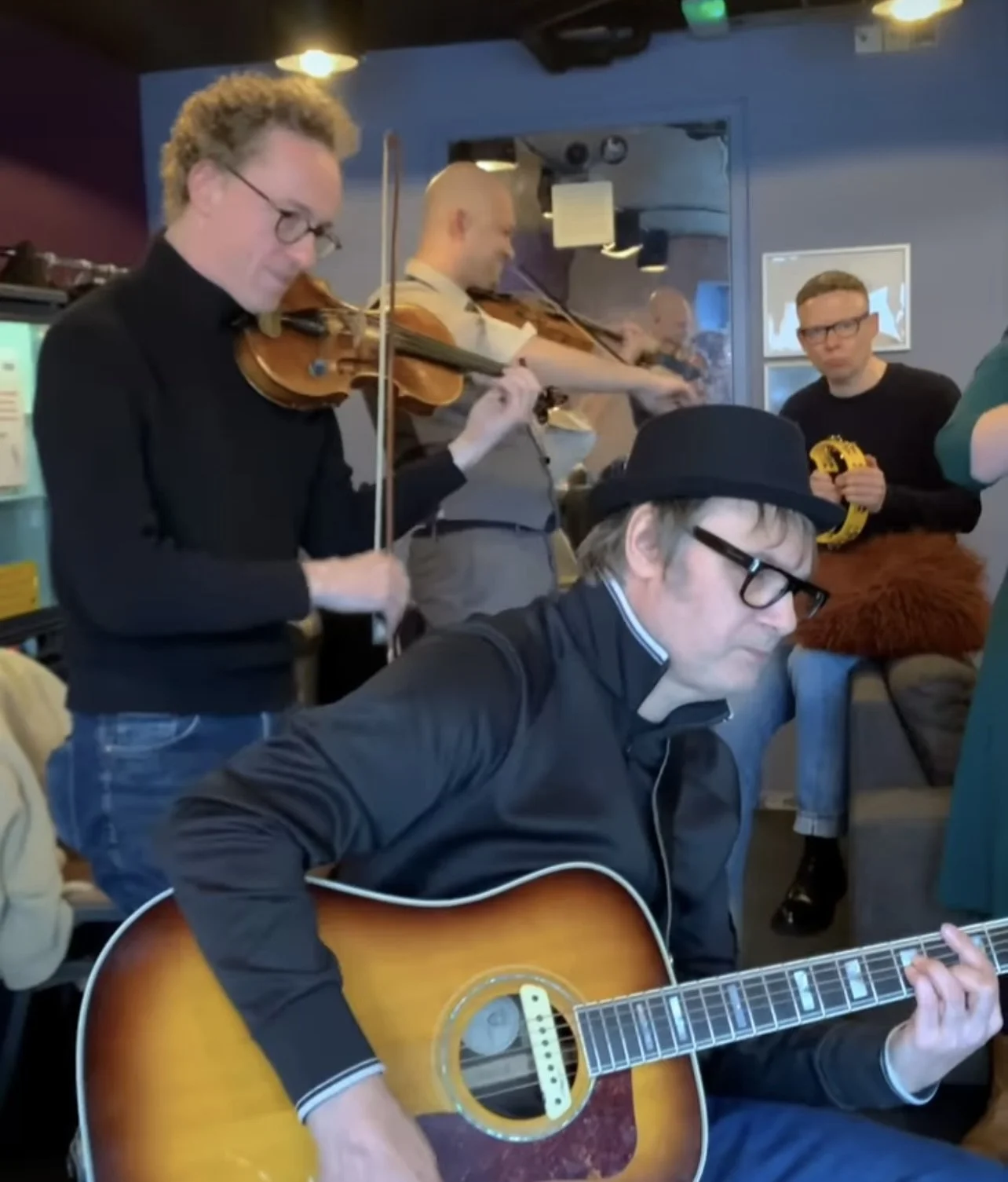 Group of musicians playing instruments, including violinists and a guitarist, in a cozy indoor setting.