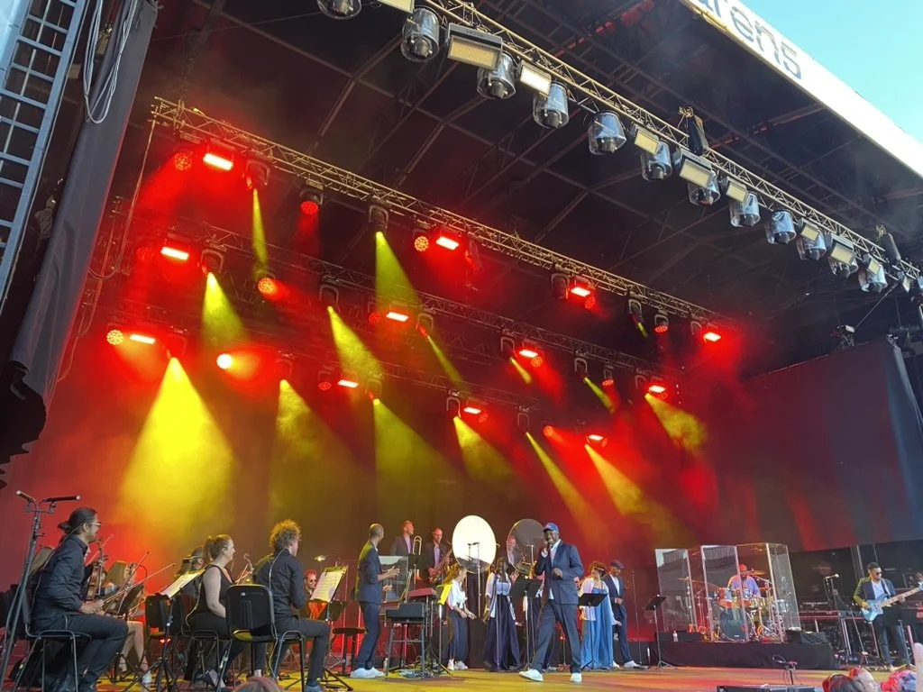 Stage with orchestra performing, lights shining in red and yellow, conductor leading, musicians playing instruments, some in masks, outside.