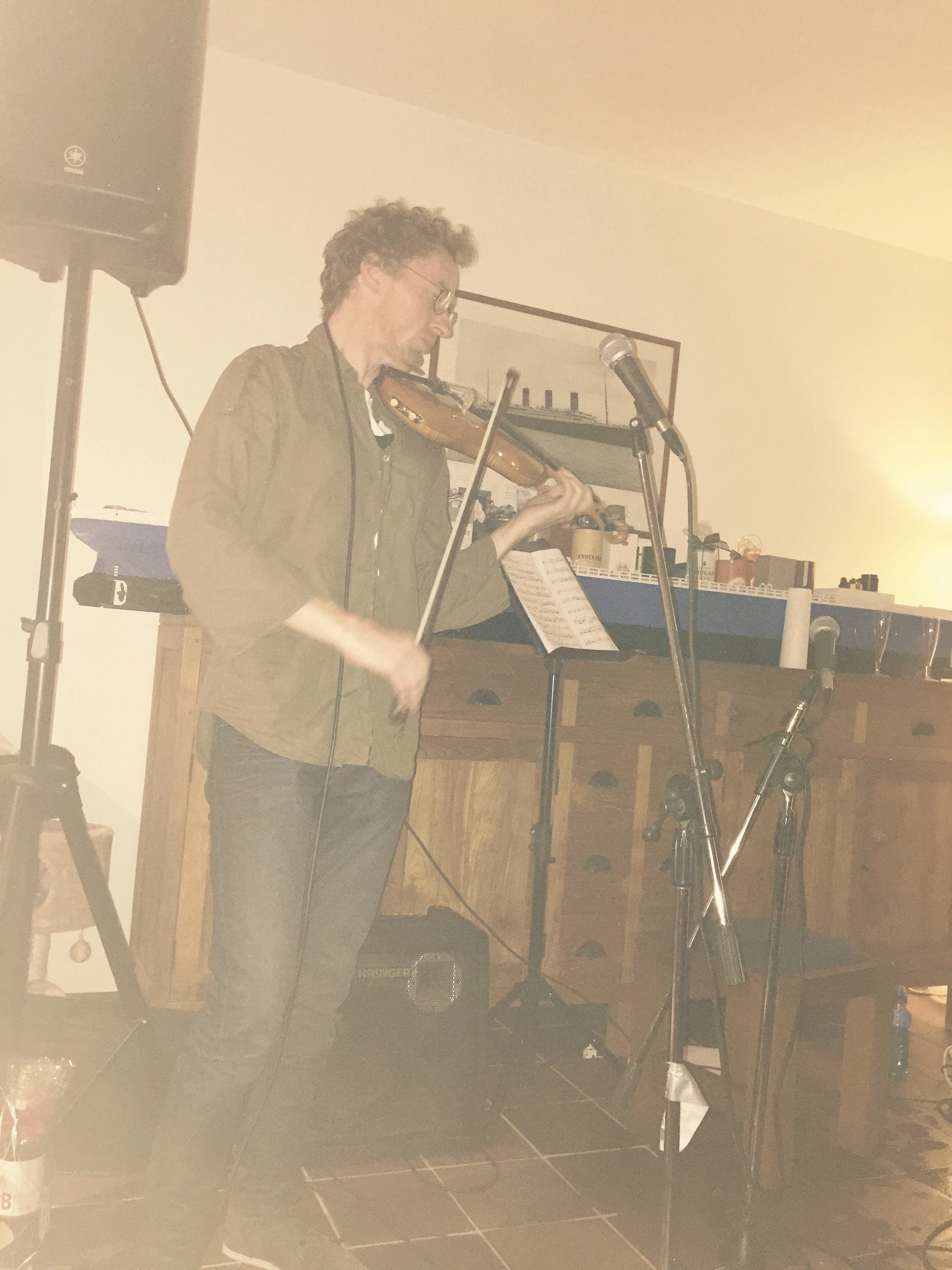 A man playing a violin indoors, standing near a microphone and music stand with sheet music. There is a large speaker on a stand to the left, and a shelf with various items in the background.