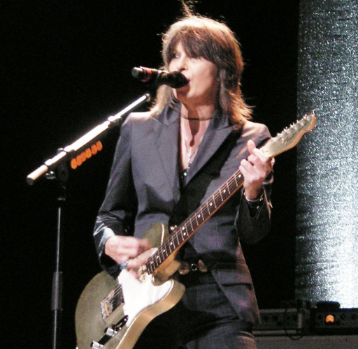 Female musician performing on stage with a microphone and electric guitar, wearing a dark blazer and black pants.