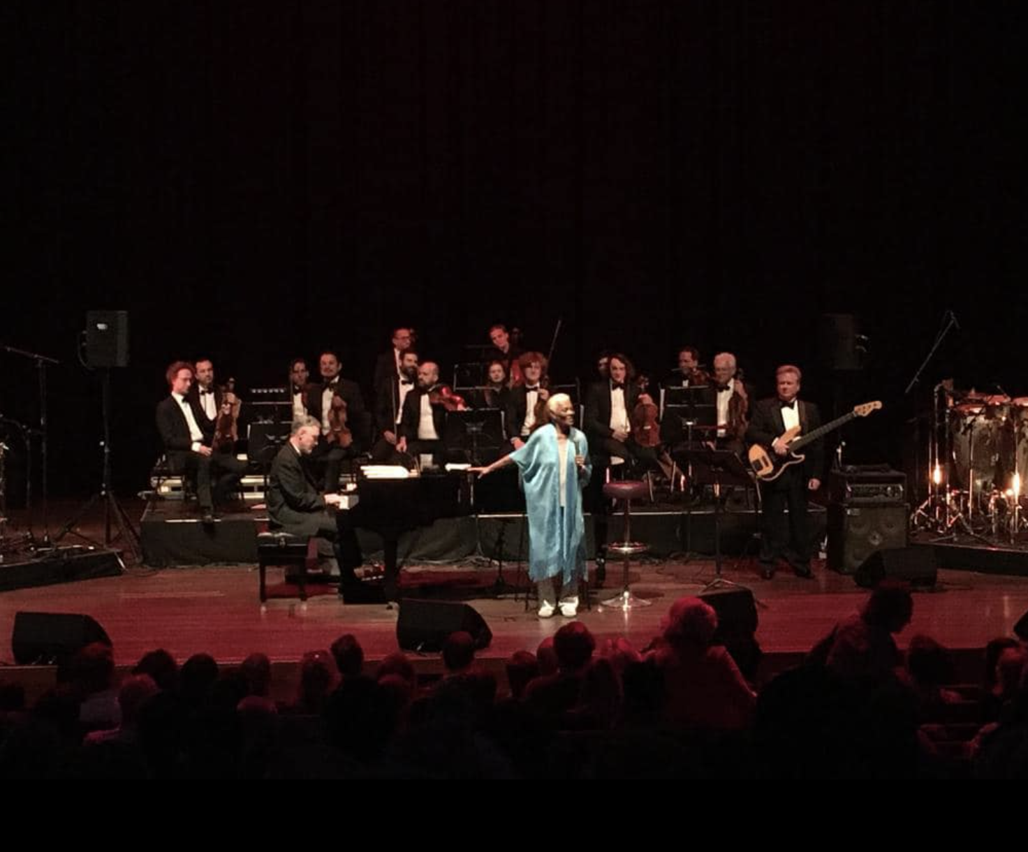 Jazz concert on stage featuring a female singer in blue attire, a pianist, and a bassist, with an orchestra in tuxedos seated behind them and audience in foreground.