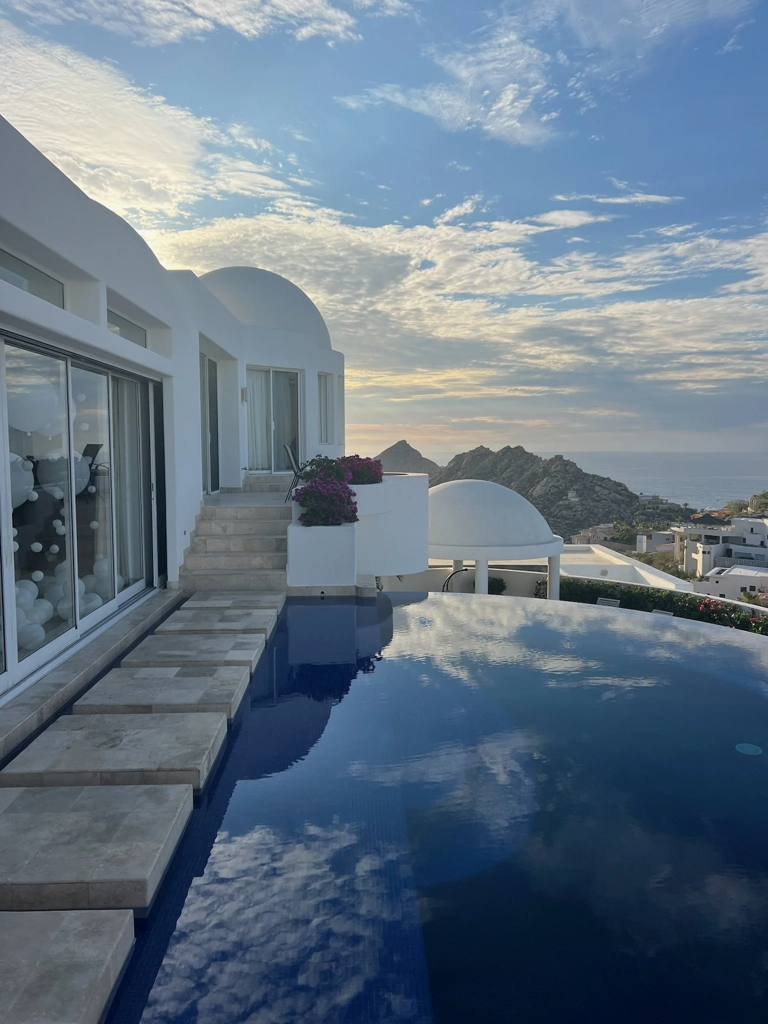 View of a modern white building with large glass doors, a swimming pool reflecting the sky, purple flowers, and distant mountains under a partly cloudy sky at sunrise or sunset.