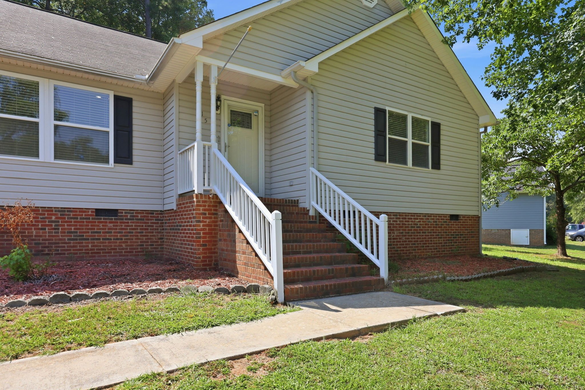 Front exterior of a Raleigh-area home with light siding, brick foundation, front steps, white railings, and a maintained lawn.