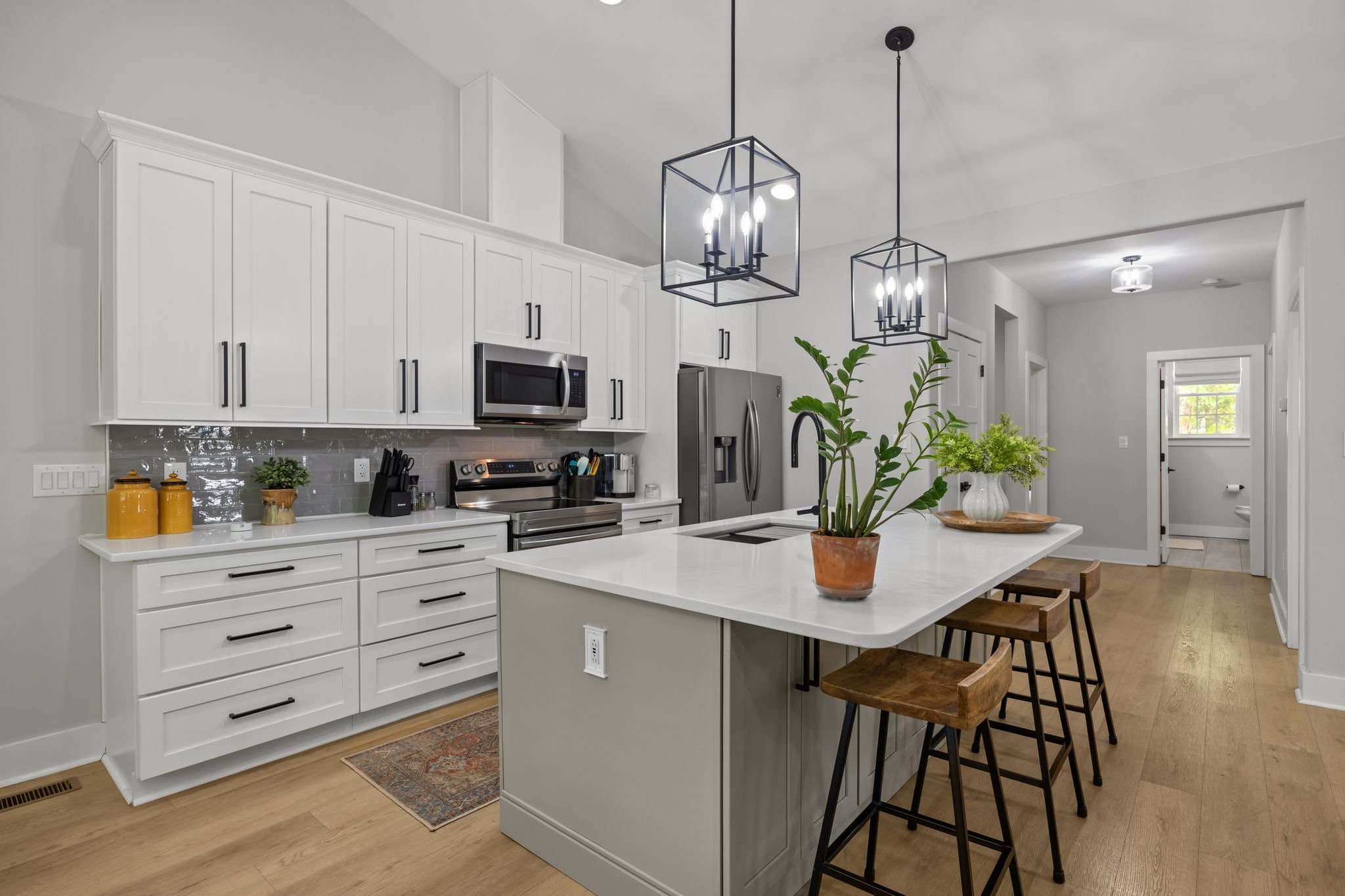 Modern kitchen with white cabinets, stainless steel appliances, black pendant lights, a kitchen island with bar stools, a potted plant, and a hallway leading to a window.