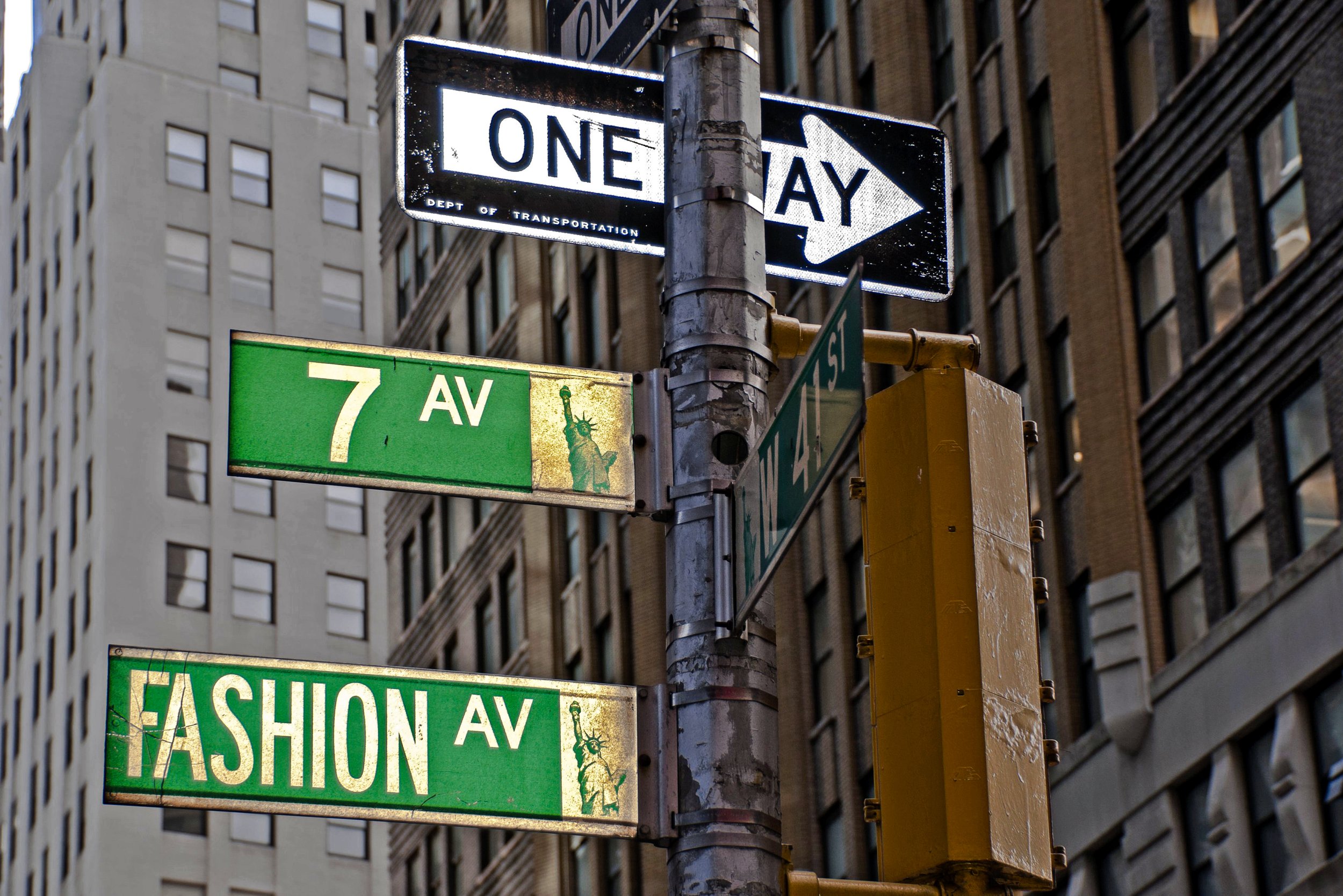 City street signs indicating one way and street names 7th Avenue and Fashion Avenue, with tall buildings in the background.