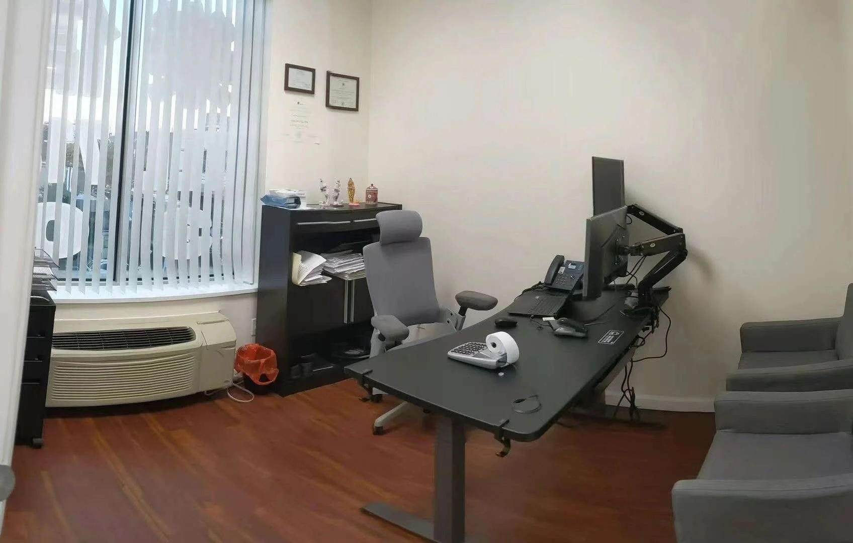 Office room with desk, two computer monitors, keyboard, telephone, grey office chair, window with vertical blinds, air conditioning unit, framed certificates on wall, and some small decorative items on a shelf.