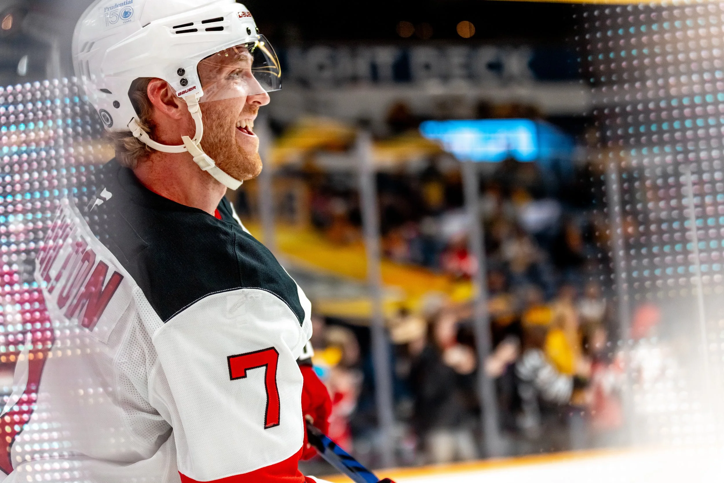 A hockey player with a beard wearing a white and red jersey and a white helmet, standing on the bench during a game. The jersey has the number 7, and the helmet features a logo and a sticker indicating 150 years of Prudential. The background is blurr