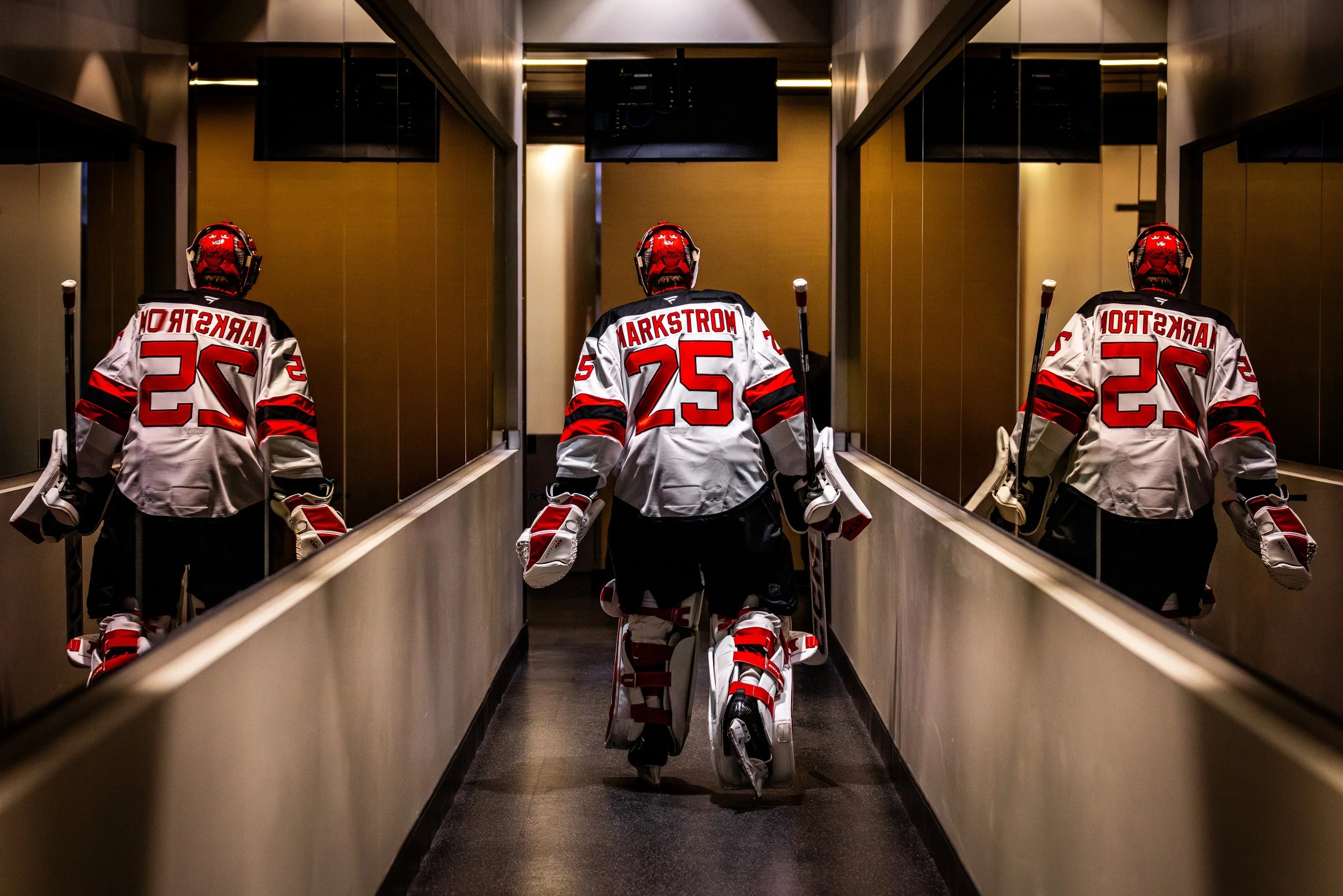 Ice hockey players in uniform and helmets push a cart with hockey gear through an indoor corridor, seen from behind.